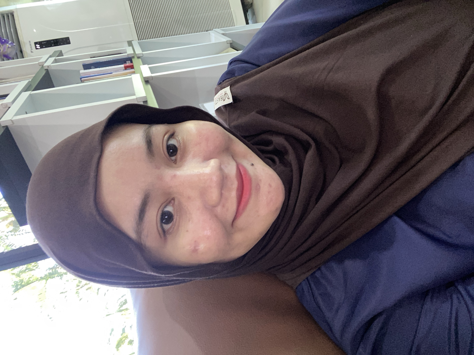 A young woman with light skin and dark hair, wearing a brown hijab and a navy blue top, is photographed from the chest up. She is looking directly at the camera and smiling with her mouth slightly open, revealing red lipstick. Her right cheek has a few visible blemishes. She is sitting in what appears to be a room with a large window in the background showing greenery. To her right, a white bookshelf with several books and an air conditioning unit can be seen. The overall impression is a casual selfie taken indoors, possibly in a home or office environment.
