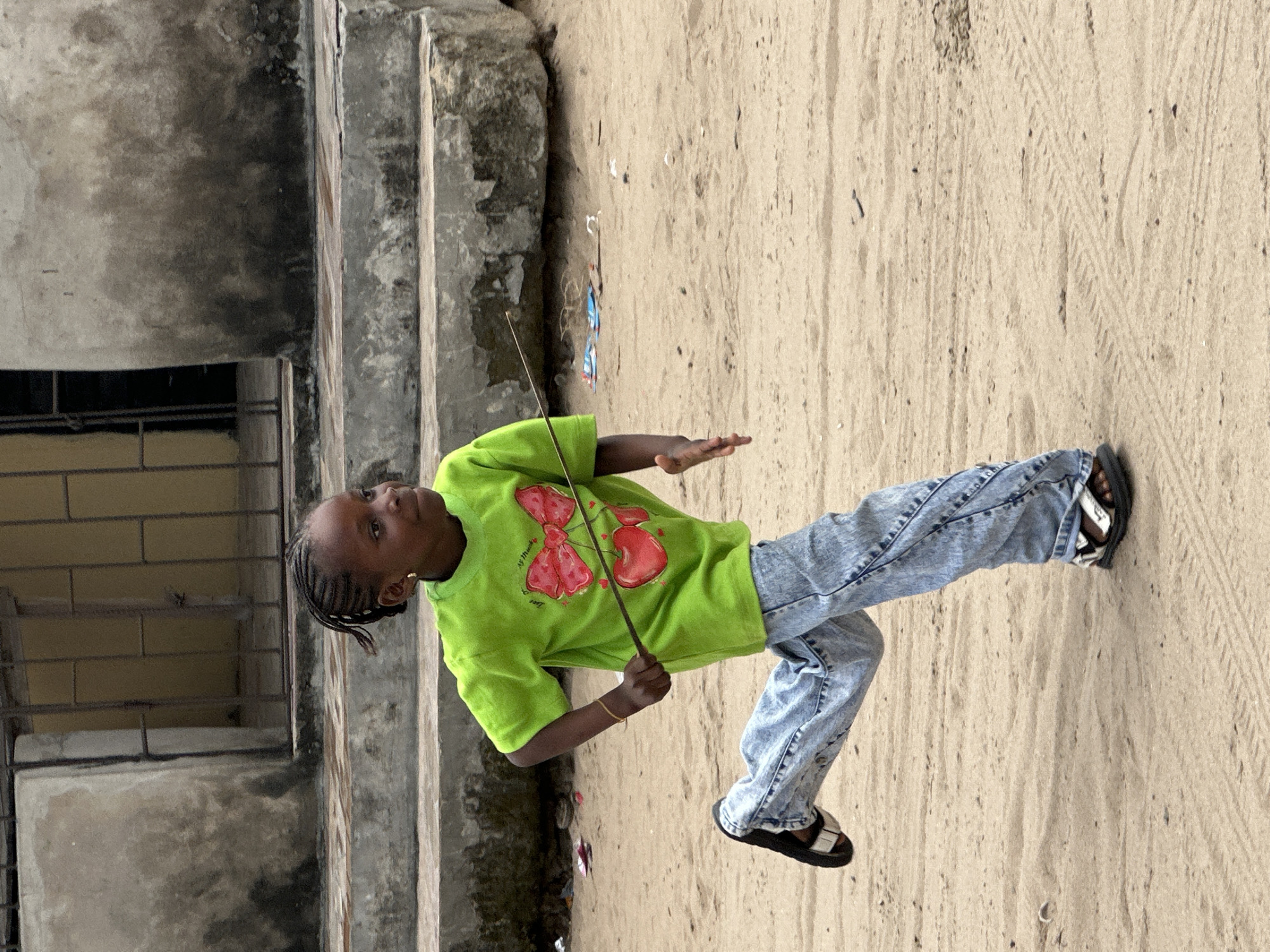 A young Black girl with braided hair is running in the sand. She is wearing a bright green t-shirt with a cherry and bow graphic on it, light blue jeans, and black sandals. She is holding a thin stick in her right hand. In the background, there are concrete stairs and a building with a barred window. The ground is sandy with some scattered debris. This scene appears to be in a residential area of Badagry, Nigeria, given the architectural style and the sandy terrain common in the coastal regions of the city.