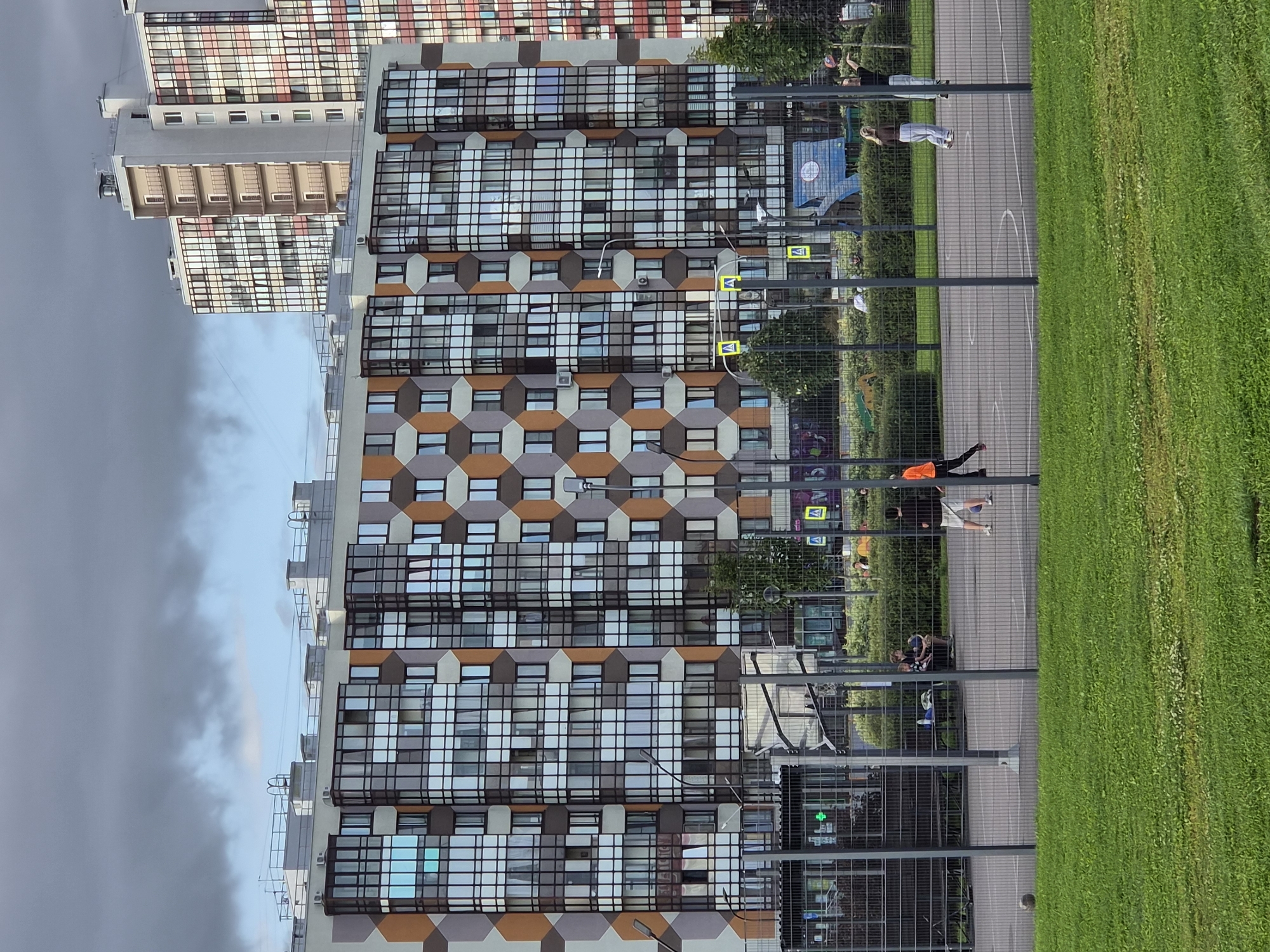 This is a medium shot, eye-level, vertical, outdoor, daytime photograph of a basketball court and residential buildings in Kudrovo, Russia. In the foreground, a well-maintained lawn is visible. Behind the lawn, a gray metal fence encloses a basketball court. On the court, three young people are playing basketball. One person in an orange shirt and white shorts is dribbling a ball, while another person in a black shirt and white shorts is about to shoot. A third person in a black shirt and black shorts is standing nearby. In the middle ground, behind the basketball court, is a dense green hedge. Behind the hedge, a modern, multi-story residential building with a geometric facade of beige, brown, and gray diamond shapes and numerous windows is prominent. To the right of this building, a taller residential building with a predominantly white facade and balconies is visible. The sky above is overcast with dark gray clouds.