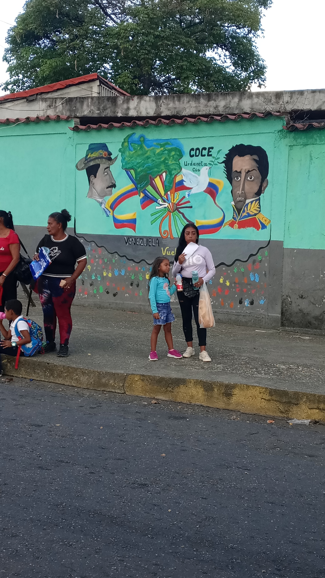 A medium shot, taken outdoors, captures a street scene with several individuals gathered on the sidewalk next to a green wall adorned with a mural. The location is in the city of Unknown, Unknown.
The mural features portraits of historical figures, the word "Venezuela," and an array of colorful handprints. A young girl in a blue top and denim shorts stands near a woman in a lilac-colored shirt and black leggings. The woman is holding a piece of paper and a plastic bag.
To the left, another woman in a black top and patterned leggings is standing with her back partially turned. A young child sits on the curb, near the woman. The street surface is dark and rough, indicating a paved road. Above the wall, green trees and a portion of a building can be seen.