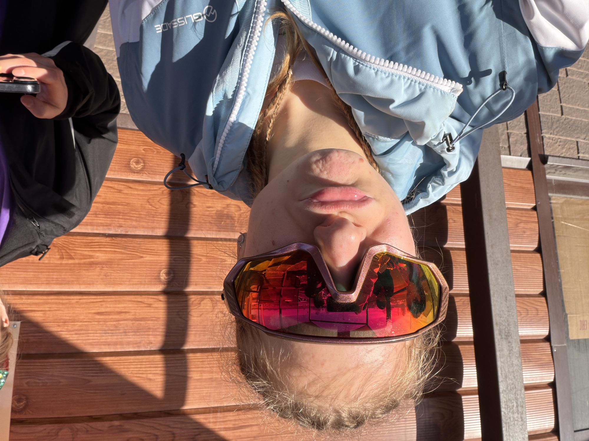 A woman with blonde hair and braids sits on a wooden bench, wearing iridescent ski goggles that reflect a cityscape. She is wearing a light blue and white jacket with "GLISSADE" visible on the left shoulder. Beside her, someone holds a smartphone. The bench is made of horizontal wooden planks. The reflection in her goggles suggests a city environment, and based on the location, this could be near Gory, Russia.