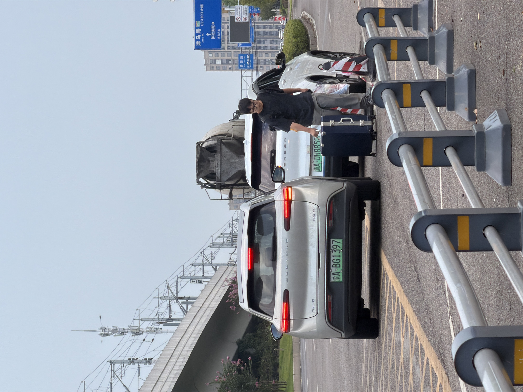 In Jiangjin, China, on a cloudy day, a silver car with a green license plate reading "渝A BG1397" is parked in the left lane of a road. A white SUV with a license plate reading "渝A DH8852" is to its right. A man wearing glasses, a black t-shirt, and gray pants is standing between the two vehicles, pulling a dark blue suitcase. He is also carrying a plastic bag in his left hand. Behind the white SUV, a cement mixer truck is visible. In the background, apartment buildings and an elevated train track with overhead wires are present. Road signs in Chinese are visible, indicating directions to "津马路" (Jinma Road) and "几江长江大桥" (Jijiang Yangtze River Bridge). The road is marked with yellow lines, and there are temporary barriers with reflective yellow stripes along the right side of the frame.