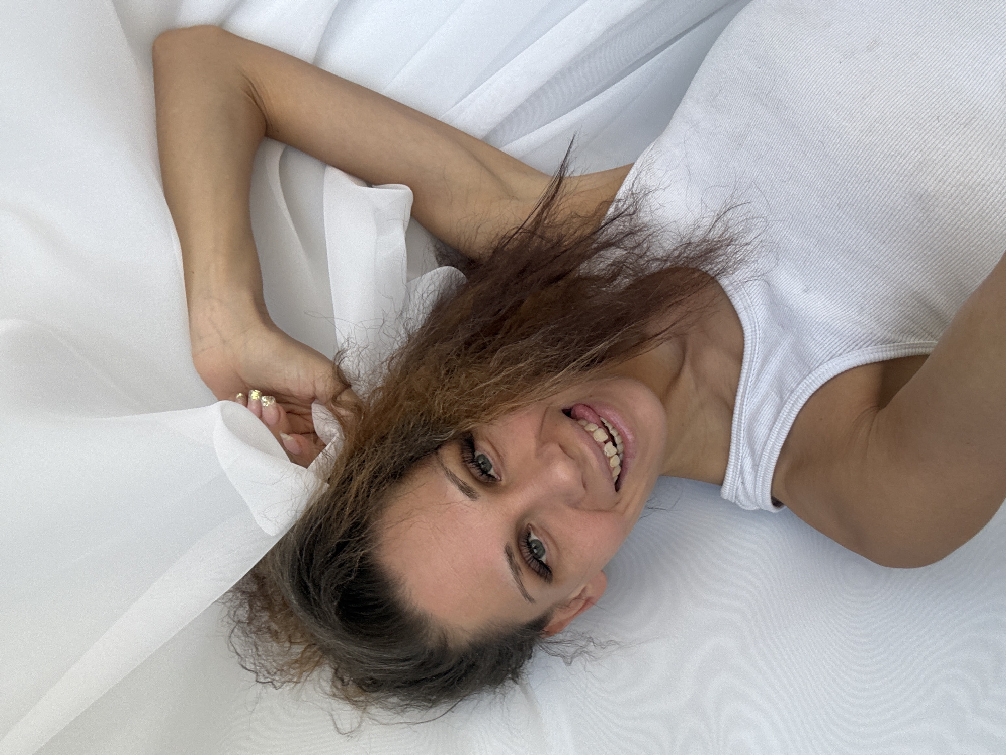An overhead shot captures a young adult woman lying on a soft, white surface, likely a bed covered with sheets and sheer fabric. Her head is tilted, and she is looking directly at the camera with a joyful, playful expression, a wide smile revealing her teeth, and her tongue slightly extended. She has green-blue eyes framed by long eyelashes and eyebrows. Her long, curly brown hair, with some lighter and possibly graying strands near the roots, spreads around her head on the white material.
She is wearing a simple white ribbed tank top. Her arms are raised above her head and resting on the white fabric. Her left hand (visible in the upper left of the frame), adorned with golden, glittery nail polish, is gently holding or resting on a fold of the sheer white fabric. The background consists entirely of this light, flowing white fabric, creating a soft and bright setting. The lighting is even and diffused, suggesting an indoor environment or soft natural light. No specific location cues are visible within the image to suggest Rusanovo, Russia, beyond the provided context. There is no visible text.