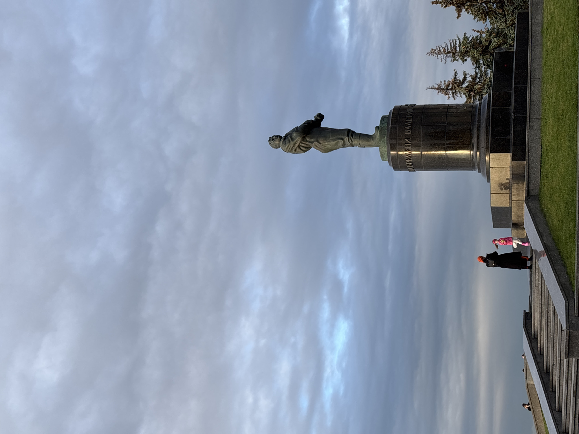 The image shows a statue of a man standing on a pedestal in a park. The statue is made of bronze and depicts a man in military uniform. The pedestal is made of dark granite and has inscriptions on it. In the foreground, there are a woman and a child standing on a staircase. The woman is wearing a black coat and an orange hat, and the child is wearing a pink jacket and white pants. The staircase is made of granite and leads up to the statue. In the background, there are trees and a cloudy sky. The statue is of Valery Chkalov, a Soviet aviator, and is located in Chkalov Stairs, Nizhny Novgorod, Russia.