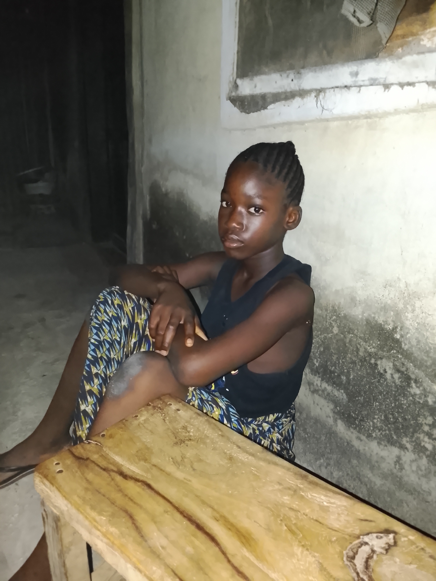 A young girl sits indoors against a stained wall in Akwanga, Nigeria. She wears a black sleeveless shirt and a patterned skirt, resting her arms on her knees. Her hair is braided back from her face. A wooden stool or bench sits in front of her. The lighting is dim. A dark doorway or hallway is visible to her left and a window frame or other architectural detail is visible in the upper right corner of the wall.