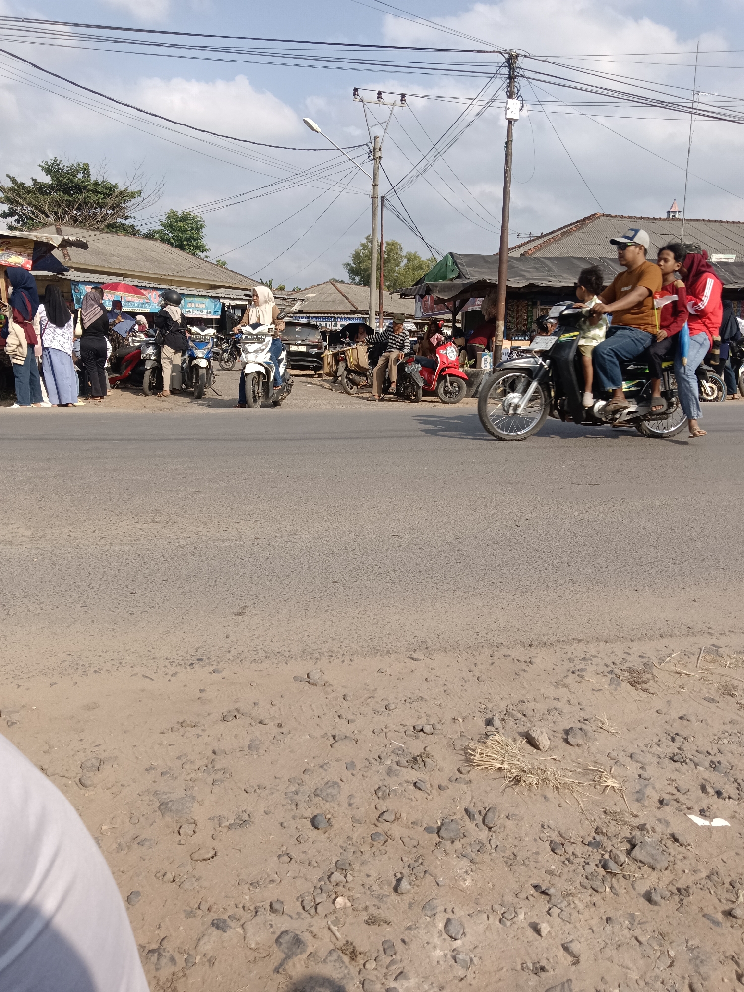 The image depicts a street scene in Tegal Gondo, Indonesia, on a sunny day with a partly cloudy sky. The foreground shows a rough, sandy roadside with scattered stones and dried vegetation. A portion of a person's white clothing is visible in the bottom left corner. The main focus of the image is a busy street with several people and motorcycles.
On the right side of the street, a man wearing a brown t-shirt and a blue baseball cap is riding a motorcycle with three passengers: a child in front, another child in the middle, and a woman wearing a red jacket behind the man. They are all looking forward, and the children appear to be holding onto the man.
To the left of the motorcycle, a white scooter is being ridden by a woman in a light-colored hijab and white pants. She is looking forward. Further to the left, there are several other motorcycles parked or being ridden. A few individuals, including women in hijabs, are standing near a row of roadside stalls. One stall has a red umbrella. There are buildings with tiled roofs in the background, some of which appear to be shops or small businesses. Power lines crisscross the sky, supported by utility poles. The overall impression is a lively, everyday scene in a rural or semi-urban Indonesian setting.