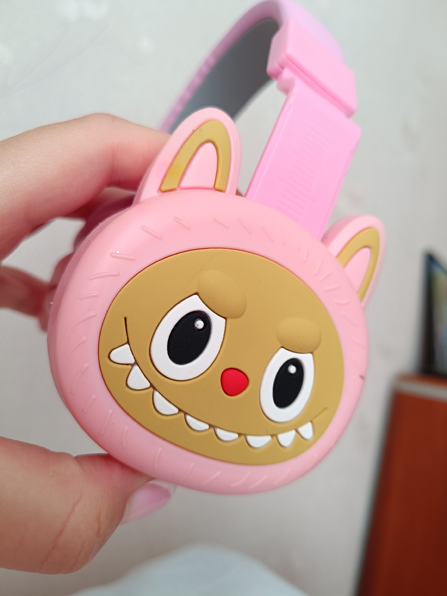 A close-up photograph depicts a pink, cartoonish pair of headphones being held in a person's hand. The headphones are designed with a monster-like face, featuring a light brown face, large white eyes with black pupils, a red nose, and white teeth. The monster is framed by a pink hood with small, rabbit-like ears edged in light brown. The headband is also pink and gray. The background is blurred and out of focus. The location is not identifiable from the provided image.