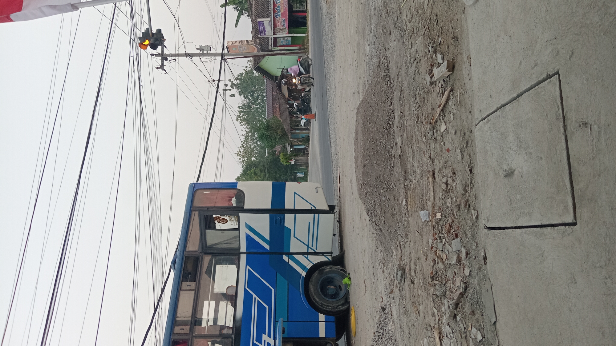 The image depicts a street scene in an outdoor, possibly urban environment. In the foreground, there is a patch of disturbed earth with gravel and scattered debris, suggesting a construction or unkempt area. Two large concrete slabs with a central seam are visible on the ground. Behind this, a blue and white bus is parked, its rear section facing the camera. The bus has visible windows and a blue stripe pattern along its side.
Further back, a road is visible with several motorcycles and riders. Some buildings with tiled roofs are present on the side of the road, with one appearing to be a shop with signage. A traffic light with an illuminated yellow signal is mounted on a pole. Overhead, a dense network of power lines crisscrosses the sky. A small pile of gravel sits on the road in the mid-ground. The sky is overcast, and the overall lighting suggests it is daytime. A sliver of a red and white flag is visible in the upper right corner of the image.