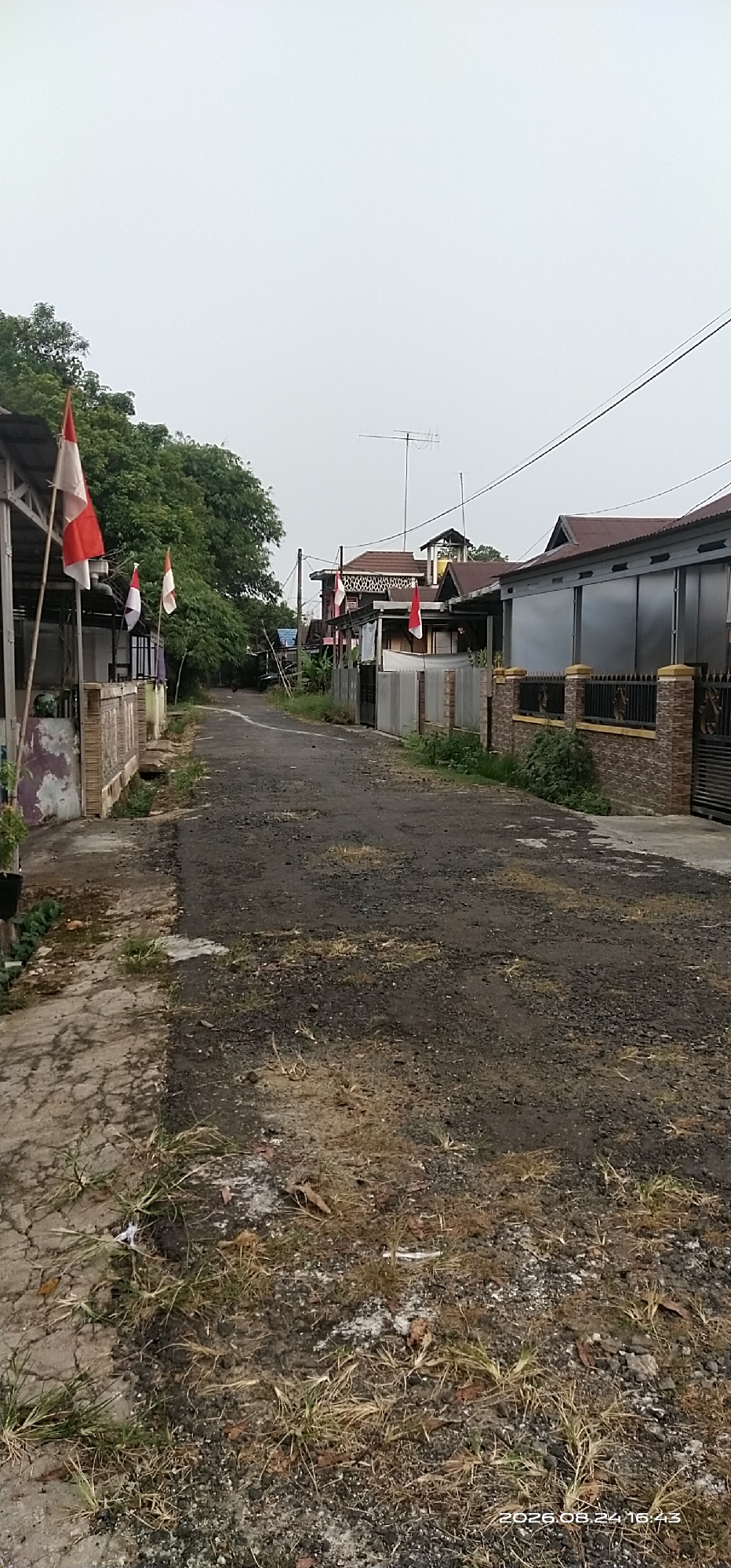 The image depicts a residential street in Matabu, Indonesia, on a cloudy day. The road is paved with asphalt and shows signs of wear, with some potholes and debris scattered on its surface. Flanking the road are houses with various architectural styles, some featuring metal roofs and brick or concrete walls. Several houses have fences made of brick and decorative metalwork. Indonesian flags, red and white, are prominently displayed on poles outside some of the residences, suggesting a national celebration or patriotic sentiment. Green trees and foliage are visible along the sides of the street and behind the houses, indicating a semi-rural or suburban setting. In the background, a television antenna is mounted on one of the rooftops. The overall scene conveys a sense of everyday life in a residential area.