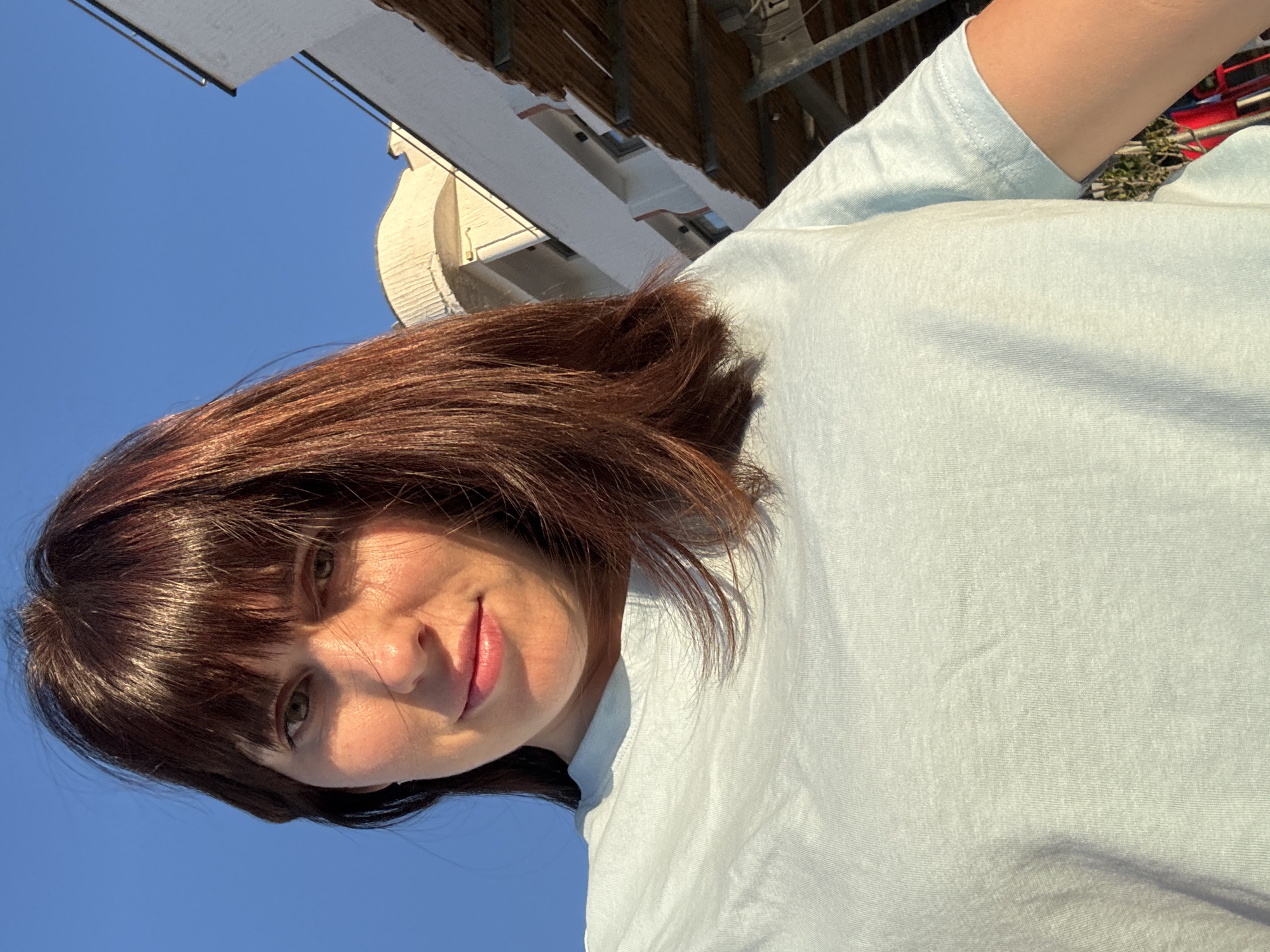 The image is a close-up, vertical selfie of a young woman with medium-length, reddish-brown hair and bangs. She is wearing a light blue t-shirt. Her face is turned towards the camera, and she has a slight smile. Sunlight illuminates her face, creating shadows and highlights. In the background, there is a clear blue sky. Above the sky, there are hints of a building with white walls and a brown roof structure, possibly an overhang or balcony, with some metal scaffolding visible. The location is Isola delle Femmine, Italy. The sunlight suggests it is daytime, likely morning or afternoon.