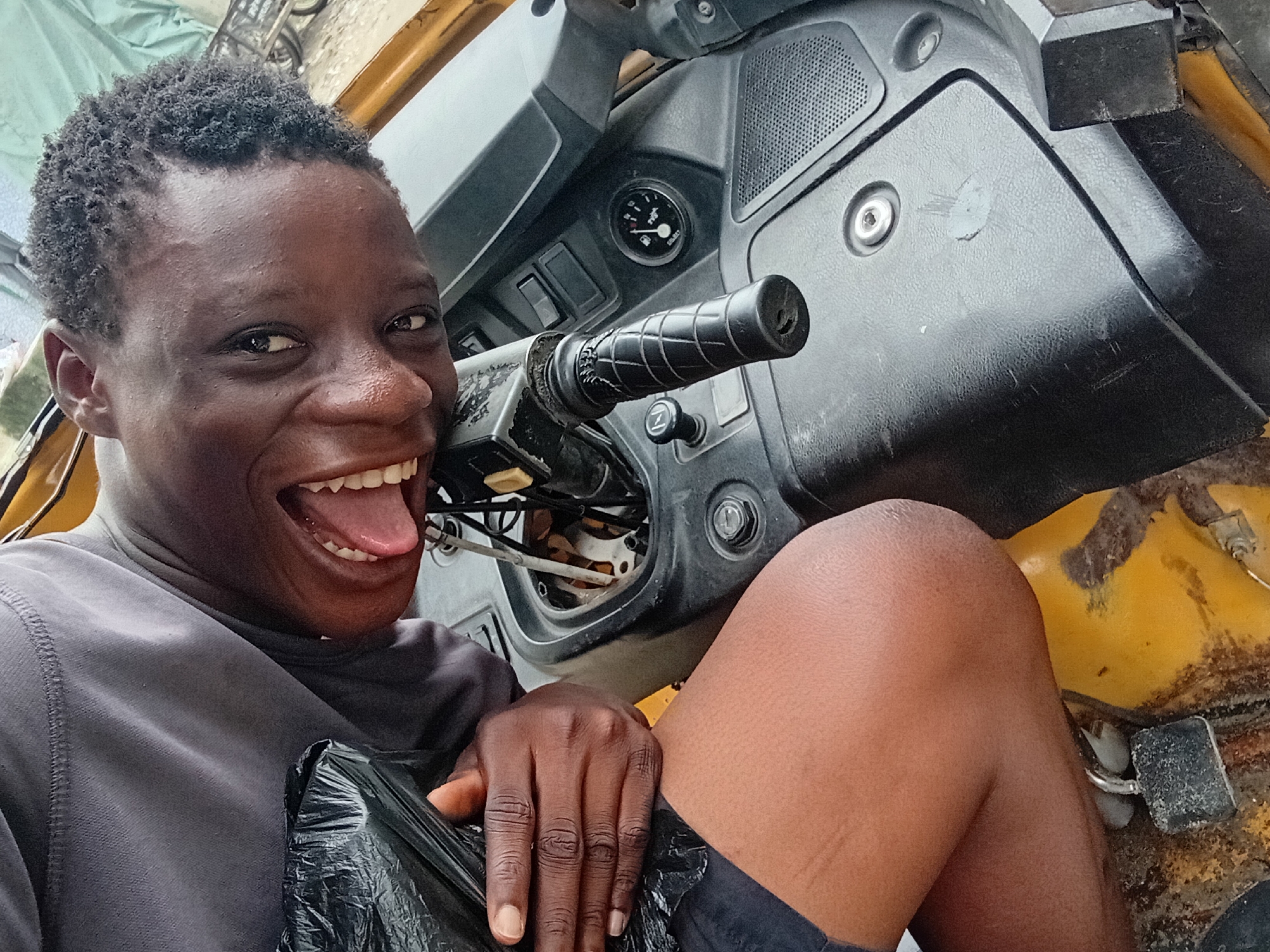 An adult is sitting in the driver's seat of a yellow three-wheeled vehicle. They are leaning towards the camera, smiling broadly with their mouth open and tongue visible. The person is holding a black plastic bag in their left hand, which is resting on their lap. They are wearing a dark gray t-shirt and dark shorts. The interior of the vehicle is visible, showing the steering wheel, dashboard with gauges and switches, and part of the floor. The environment appears to be outdoors, possibly on a street, as indicated by the light and the presence of other elements in the background which are blurred.