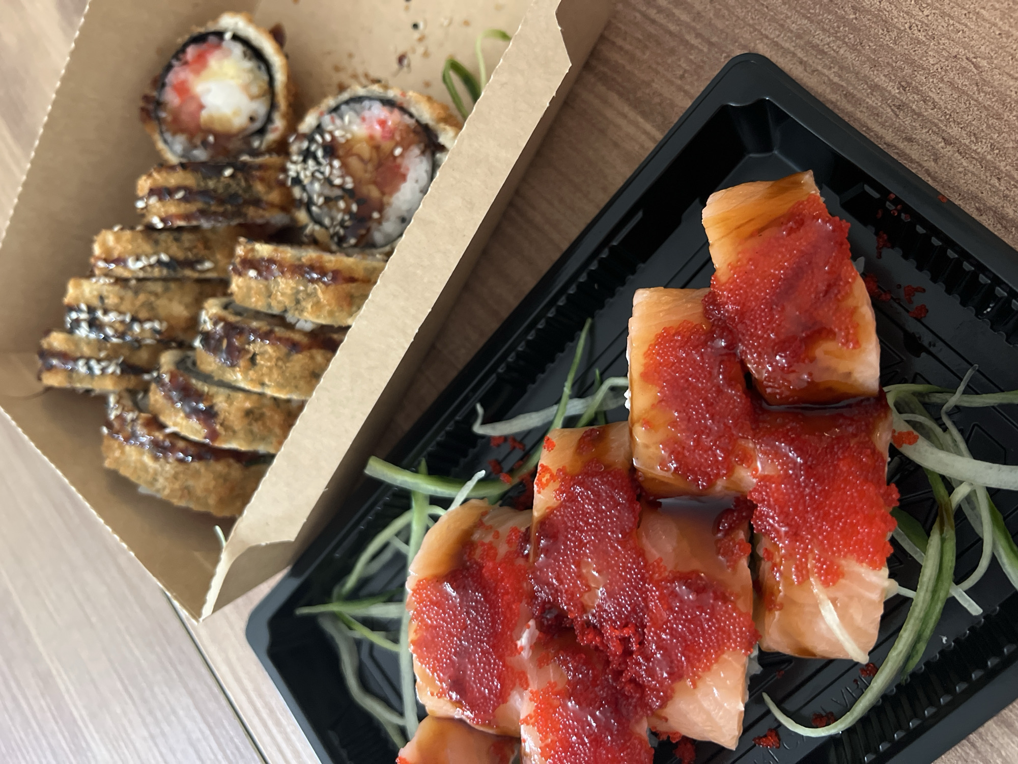On a wooden surface, there are two containers of sushi. The left container is brown and contains fried sushi rolls with sesame seeds and a drizzled sauce. The right container is black and contains sushi rolls topped with salmon and red roe. There are also some green onions in the right container. The location in Budva, Montenegro is not discernable from this image.