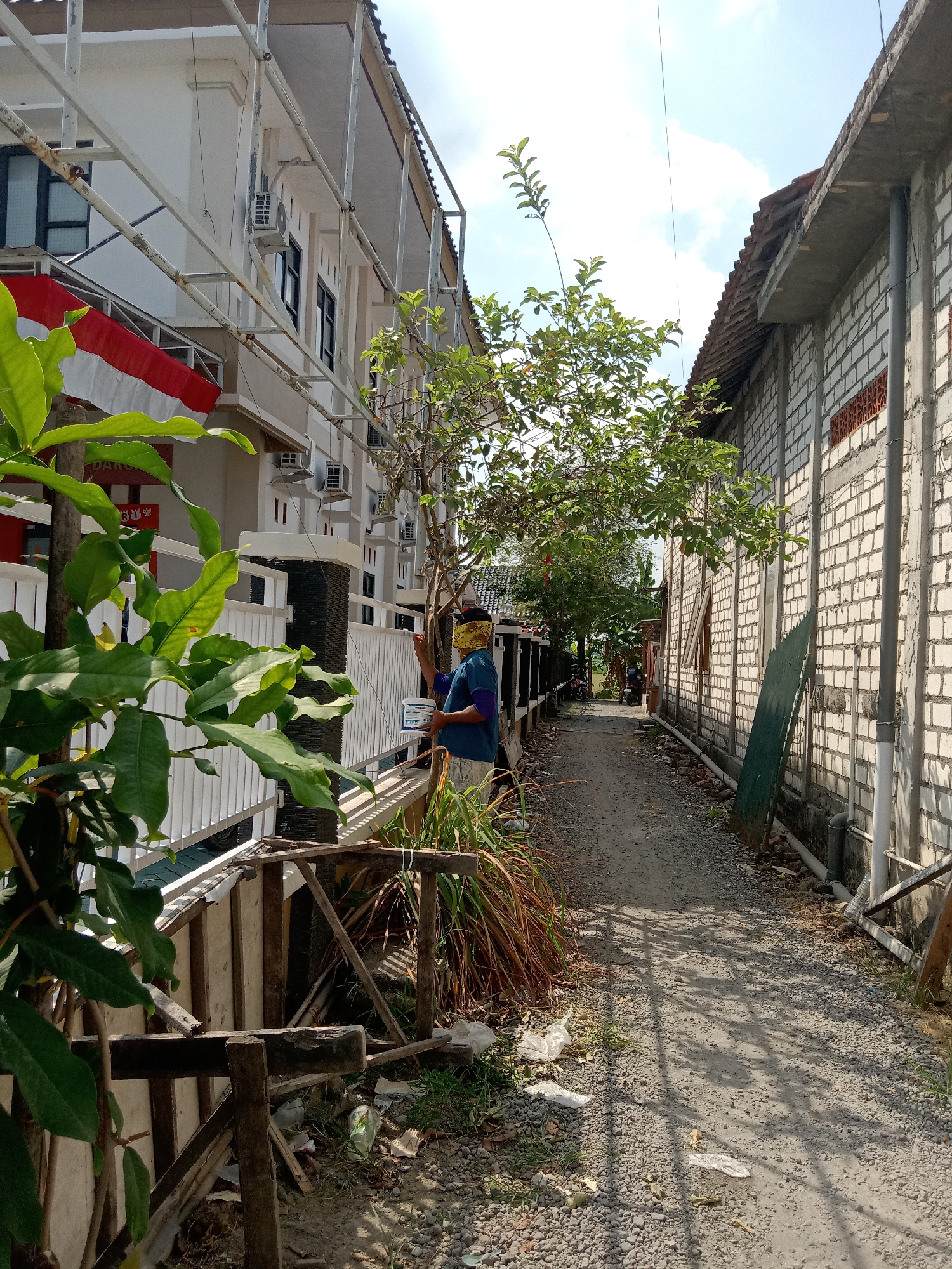 A person wearing a blue shirt and a yellow bandana is standing on a gravel path in an alleyway between two buildings. The person is holding a white paint can and a brush, and appears to be painting a white railing. The building on the left is a multi-story white building with windows and an air conditioning unit. A red and white flag is visible on the building. The building on the right is made of white bricks. There are trees and vegetation on the left side of the alley. The sky is blue with some clouds. The scene appears to be in a residential area in Klepek, Indonesia.