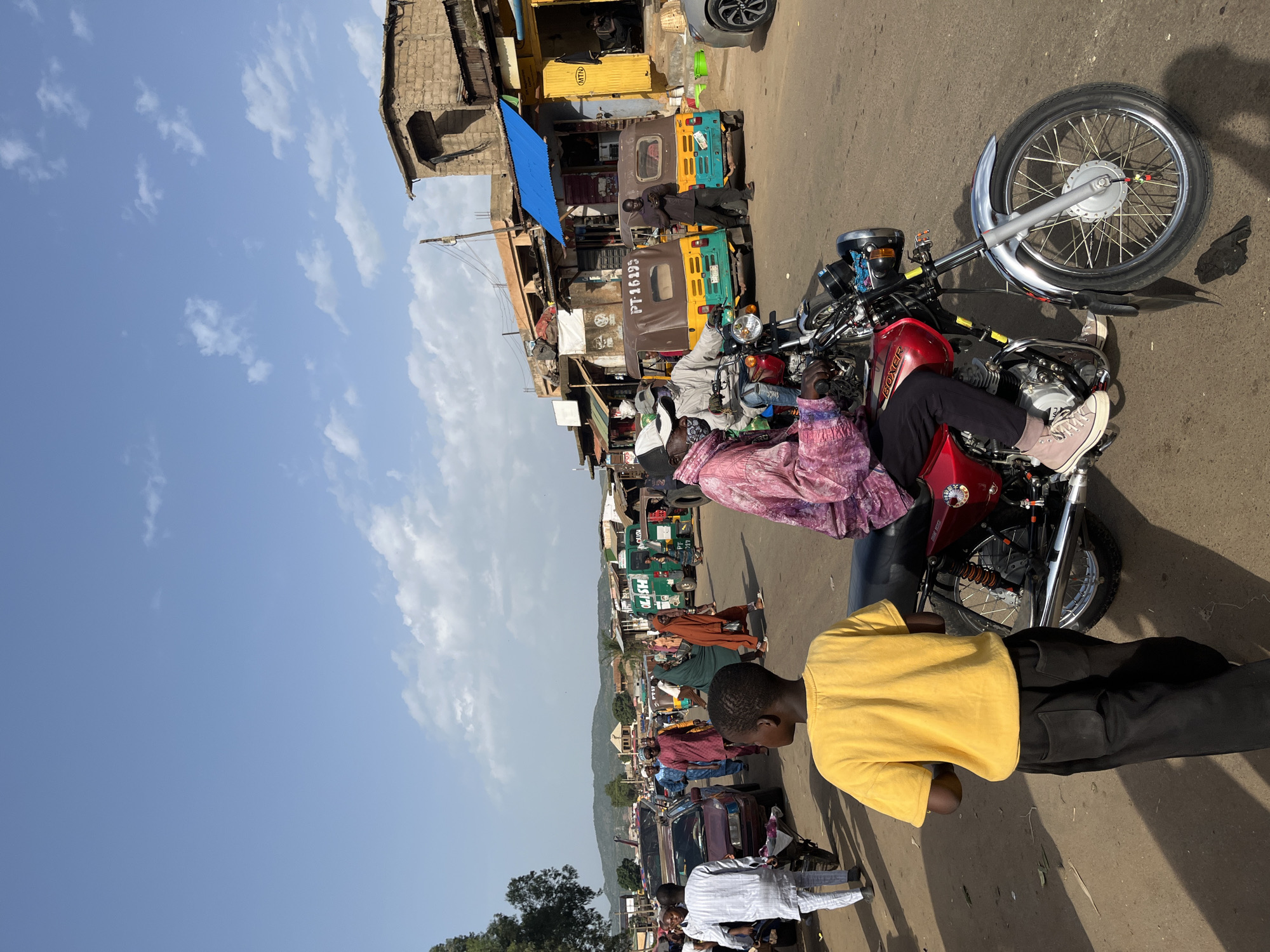 The image depicts a bustling street scene in Rafin Par, Nigeria. The foreground is dominated by a red motorcycle, with its front wheel and handlebars clearly visible. A person wearing a pink patterned shirt and dark pants, with light-colored sneakers, is seated on the motorcycle. To the right of the motorcycle, another person, a young boy wearing a bright yellow t-shirt and black pants, is also visible, facing away from the camera.
The background reveals a street lined with buildings, likely shops or market stalls, some with blue and yellow awnings. Several motorbikes and auto-rickshaws, also known as "keke napeps," are parked or moving along the street. One auto-rickshaw has the text "PT-16398" visible on its side. Another auto-rickshaw has "TOYOTA" written on it. People are walking and standing around, engaged in various activities, suggesting a lively market or commercial area. The setting appears to be outdoors during the daytime, with a clear blue sky and scattered clouds. The overall atmosphere suggests a busy, everyday scene in a Nigerian town. The lighting indicates it is likely midday or early afternoon.