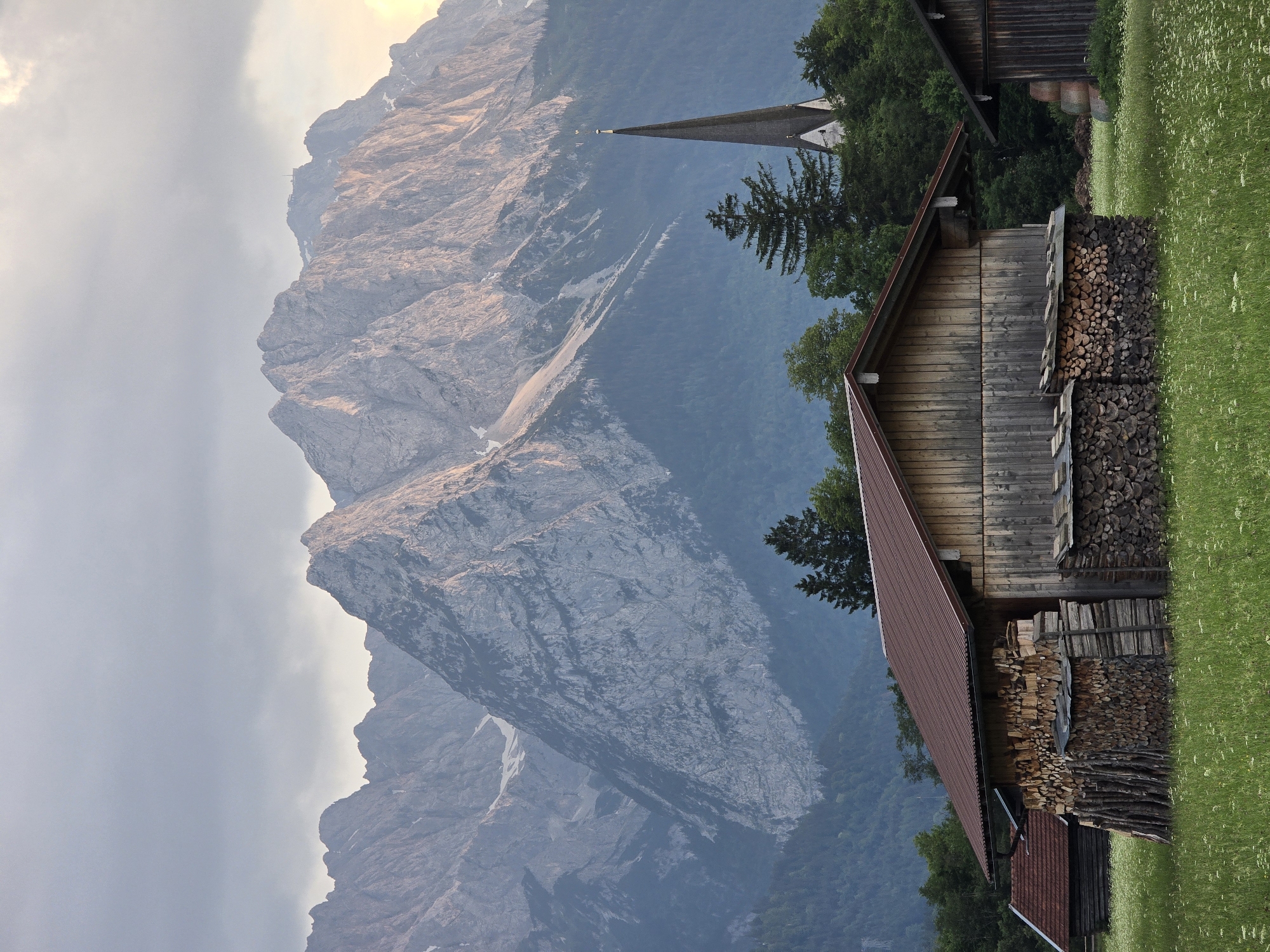 This image captures a scenic view in Garmisch-Partenkirchen, Germany, featuring a traditional wooden barn in the foreground, stacked with firewood. Beyond the barn, a meadow with scattered white wildflowers stretches towards a dense forest. In the middle ground, a church steeple rises above the trees, with a magnificent mountain range dominating the background. The mountains, partially lit by the sun, display rugged, rocky formations and patches of snow. The sky is overcast with soft, diffused light, contributing to a serene atmosphere. There are no people or animals visible in the image.