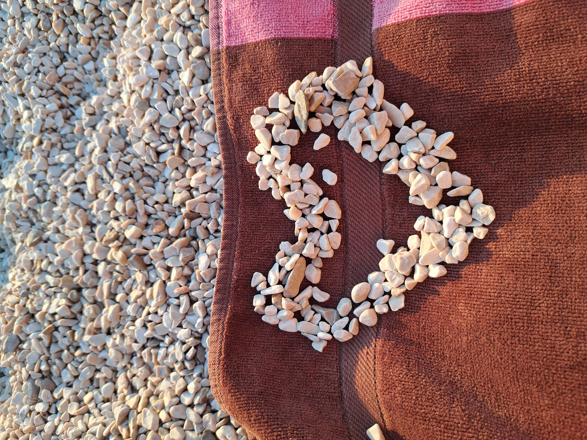 A heart shape made of white and tan pebbles is laid out on a dark brown terrycloth towel. The towel has a vertical pink stripe on the right edge. Behind the towel, a large pile of similar pebbles fills the top portion of the frame, suggesting a beach or gravelly area. The lighting suggests it is late afternoon or early morning, with long shadows cast by the pebbles. There are no people or animals visible in the image.
