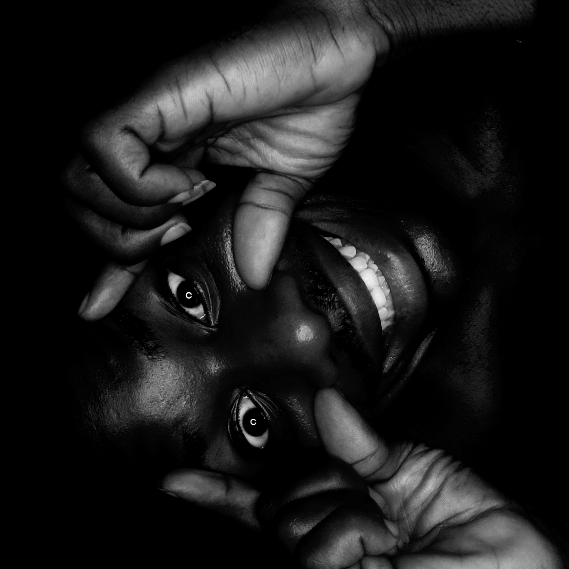 A close-up, black and white photograph depicts a person's face, seemingly upside down. Two hands frame the face, with fingers touching the skin near the eyes and nose. The person's eyes are open, with visible reflections. The mouth is open in a possible smile, revealing teeth. The background is black and indistinct. Location in Narayi, Nigeria, is not discernible from the visual information.