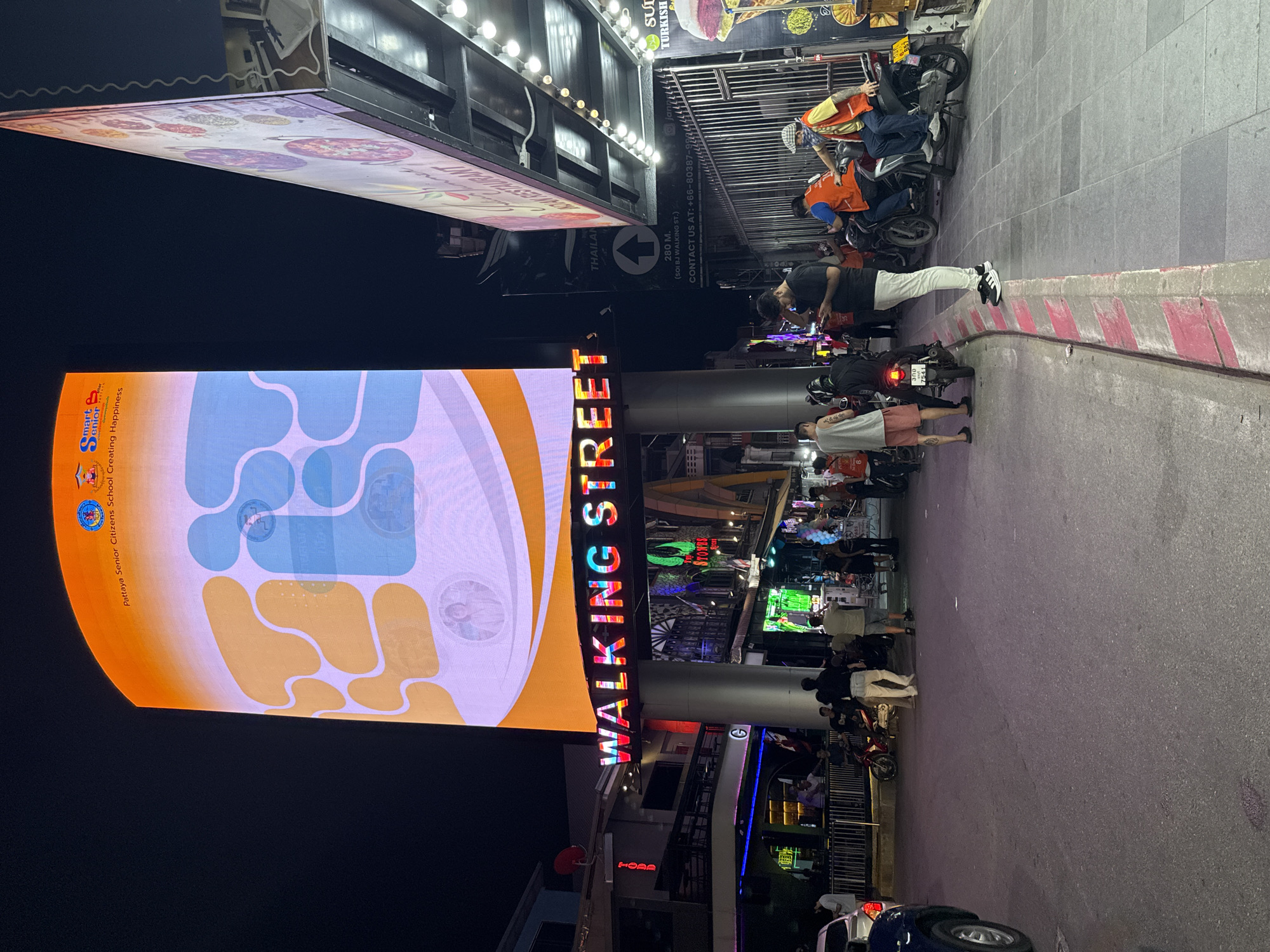 The image displays a lively night scene on a bustling street in Pattaya City, Thailand. A prominent, vertical LED display on the left side of the frame features abstract blue and orange patterns, along with the text "Pattaya Senior Citizens School Creating Happiness" and a "Smart Senior" logo. Below it, a large, multi-colored neon sign clearly spells "WALKING STREET," indicating the location.
The street is illuminated by artificial lights, revealing various people walking, standing, and interacting. Numerous motorcycles are parked along the sidewalk and curb, with individuals gathered near them. In the upper right, men wearing orange vests are visible near some of these motorcycles.
The setting is urban, characterized by buildings lining the street that house businesses, indicated by their illuminated signs. Visible text includes "SULTAN TURKISH" on a sign in the background, suggesting a restaurant. Another sign provides directional information, reading "280 M. (SOI BJ WALKING ST.)," and contact details: "CONTACT US AT: +66-80387-9984." The word "THAILAND" is also discernible on a sign. The overall scene captures the vibrant energy of a popular night entertainment and commercial district.