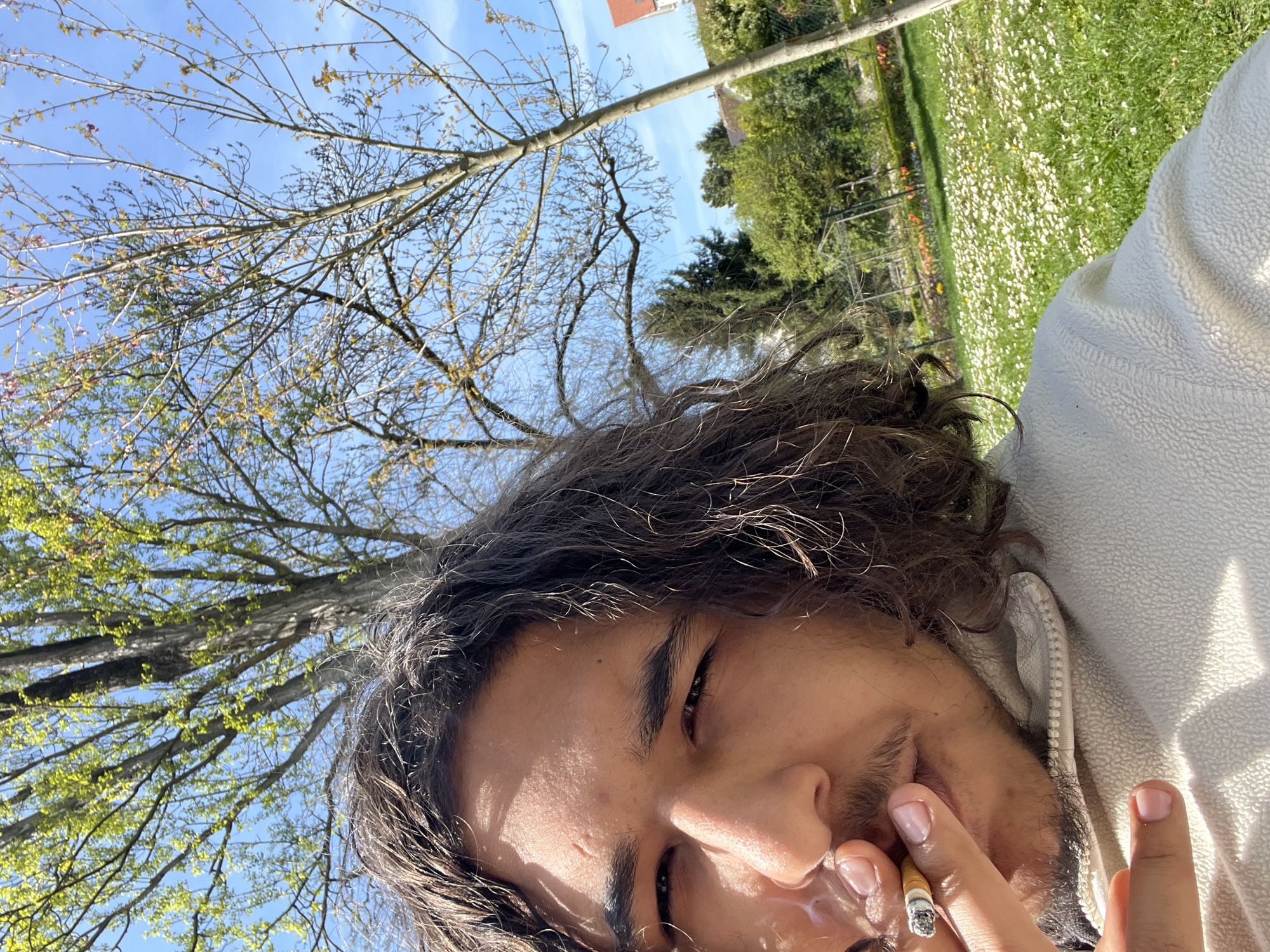 A man is shown from a low angle, seemingly reclining in a park-like setting. The background includes a grassy area covered in white flowers. Trees are visible, with branches extending into a blue sky. The man is holding a lit cigarette. The location is identified as Schiltigheim, France, based on visual cues present in the image. The man has long, dark, curly hair and is wearing a white jacket or sweatshirt.