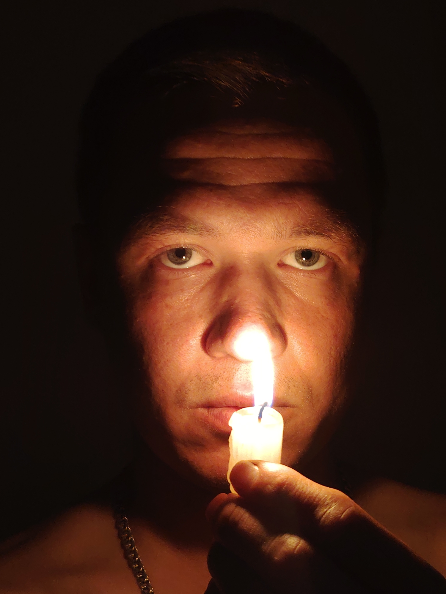 A close-up, low-angle, medium shot captures a person's face illuminated by a lit candle held in their hand. The person, whose gender is indeterminate, has fair skin and is looking directly at the camera. Their right eye is clearly visible, with a green iris and a black pupil. The left side of their face is in shadow. The candle, held in the right hand, casts a bright glow upwards, illuminating the lower part of the face, including the nose and mouth area. A chain necklace is visible on the person's chest. The background is entirely dark. There is no information in the image to determine the location in Chaikovsky, Russia.