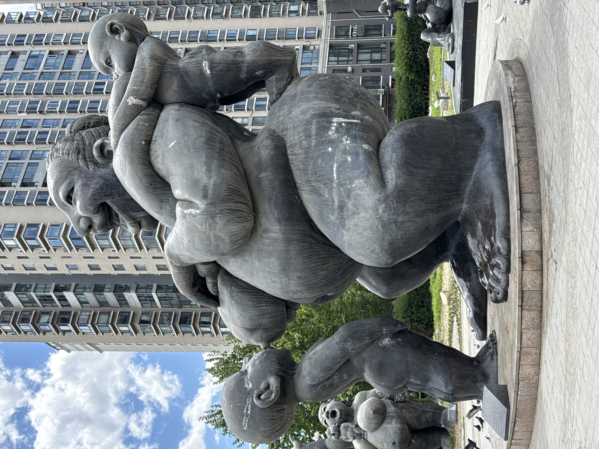 The image shows a bronze sculpture of people piled on top of each other. One person is on the ground, with other people sitting and climbing on them. In the background is a tall building with rows of windows. The sculpture is located in Changping District, China.