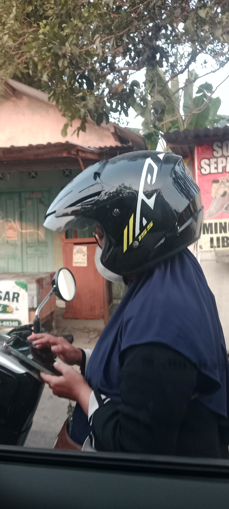 A person wearing a black motorcycle helmet with white and yellow accents is on a motorcycle in what appears to be Banyuraden, Indonesia. The person is wearing a blue hijab and a black outer garment with white accents. Their hands are on a tablet device. A motorcycle mirror is visible to the left of the person. In the background, there is a building with green doors and a tiled roof, and a sign that reads "SO SEPA". Trees and foliage are present above the building. The image is taken from a low angle, possibly from another vehicle.