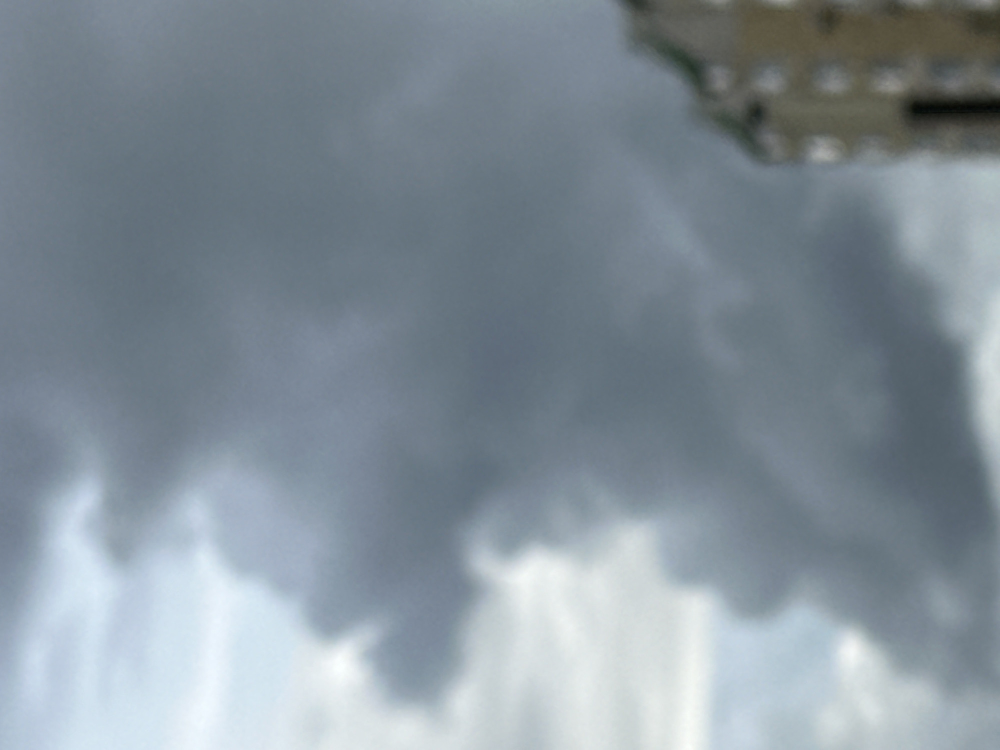 The image shows a blurry outdoor scene with a dominant cloudy sky. Dark gray and lighter gray clouds fill most of the frame, with hints of blue sky peeking through on the left. On the right side of the image, the top portion of a building with a light-colored facade and a green roof is visible. The building appears to have multiple windows. The overall impression is of an overcast or impending stormy day. The provided crops offer closer views of the clouds and the building.