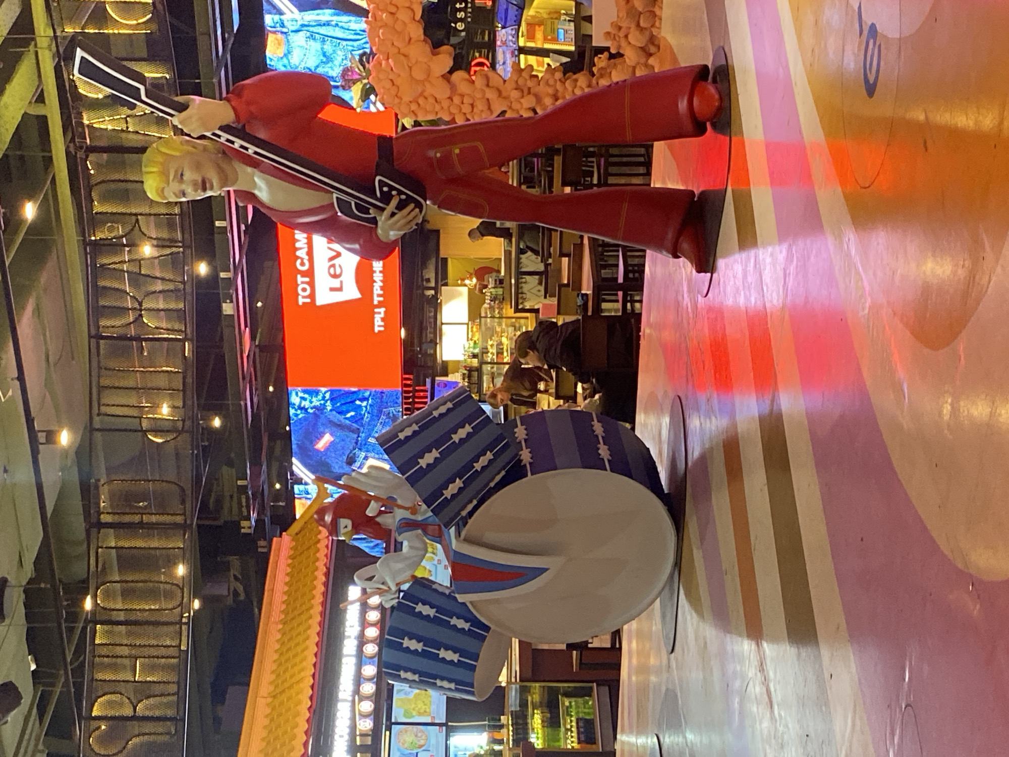 The image shows a sculpture of a rock band performing in a shopping mall. The band consists of a guitarist dressed in a red suit and a drummer. The drums are decorated with blue and white zig-zag patterns. A Levi's advertisement appears on a large screen in the background. The floor is painted with broad stripes of pink, yellow, and white. The location is likely the Passage shopping mall in Yekaterinburg, Russia.