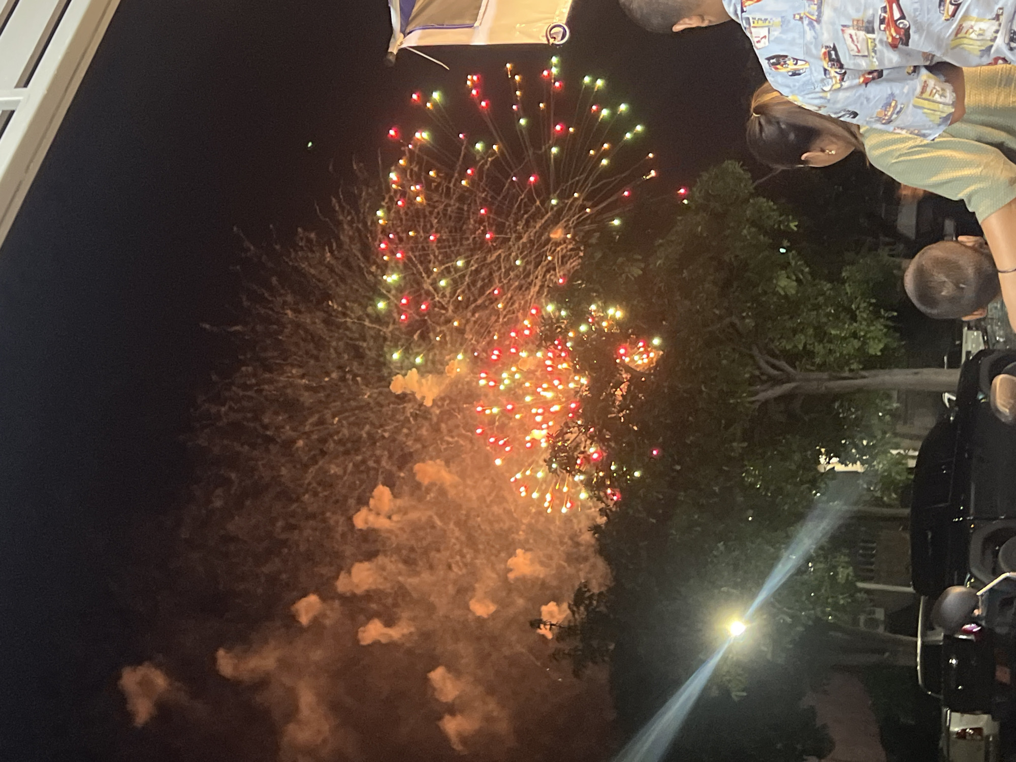 This image captures a night scene in Koh Phangan, Thailand, with fireworks illuminating the sky above a residential or commercial area. The fireworks display features bursts of red, green, and yellow lights, with orange smoke billowing in the upper left portion of the frame. In the foreground, several people are visible, including a woman in a green shirt with her back to the camera, a man in a light blue patterned shirt, and a young child with a shaved head. They appear to be watching the fireworks. In the background, trees, a street light casting a strong beam, and several vehicles, including a black pickup truck and a motorcycle, are present. The overall impression is of a festive outdoor event.