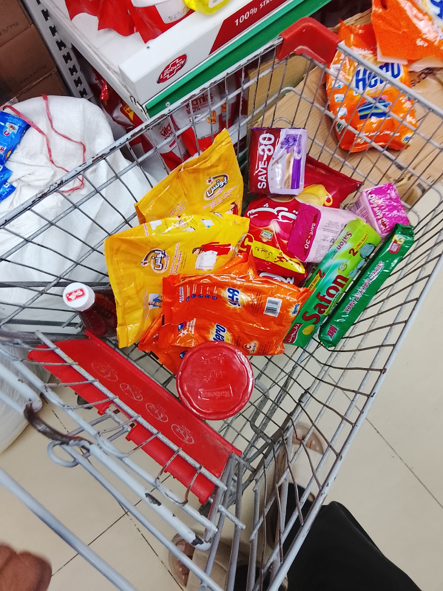 A shopping cart, presumably in a grocery store within Gulgasht Colony, Pakistan, contains various household and personal care items. Yellow packets of "Bonus" detergent are prominent, along with orange "Hero" washing powder packets. Two green packages of "Safon" dishwashing soap and a red container of "Danishol" are also visible. A "Save 30" package of what appears to be personal care wipes and a bottle of "Lifebuoy" body wash are present. A person wearing black pants and beige shoes stands behind the cart. The items are placed inside a wire shopping cart with a red child seat. The background shows shelves stocked with various products, suggesting the location is a retail environment.