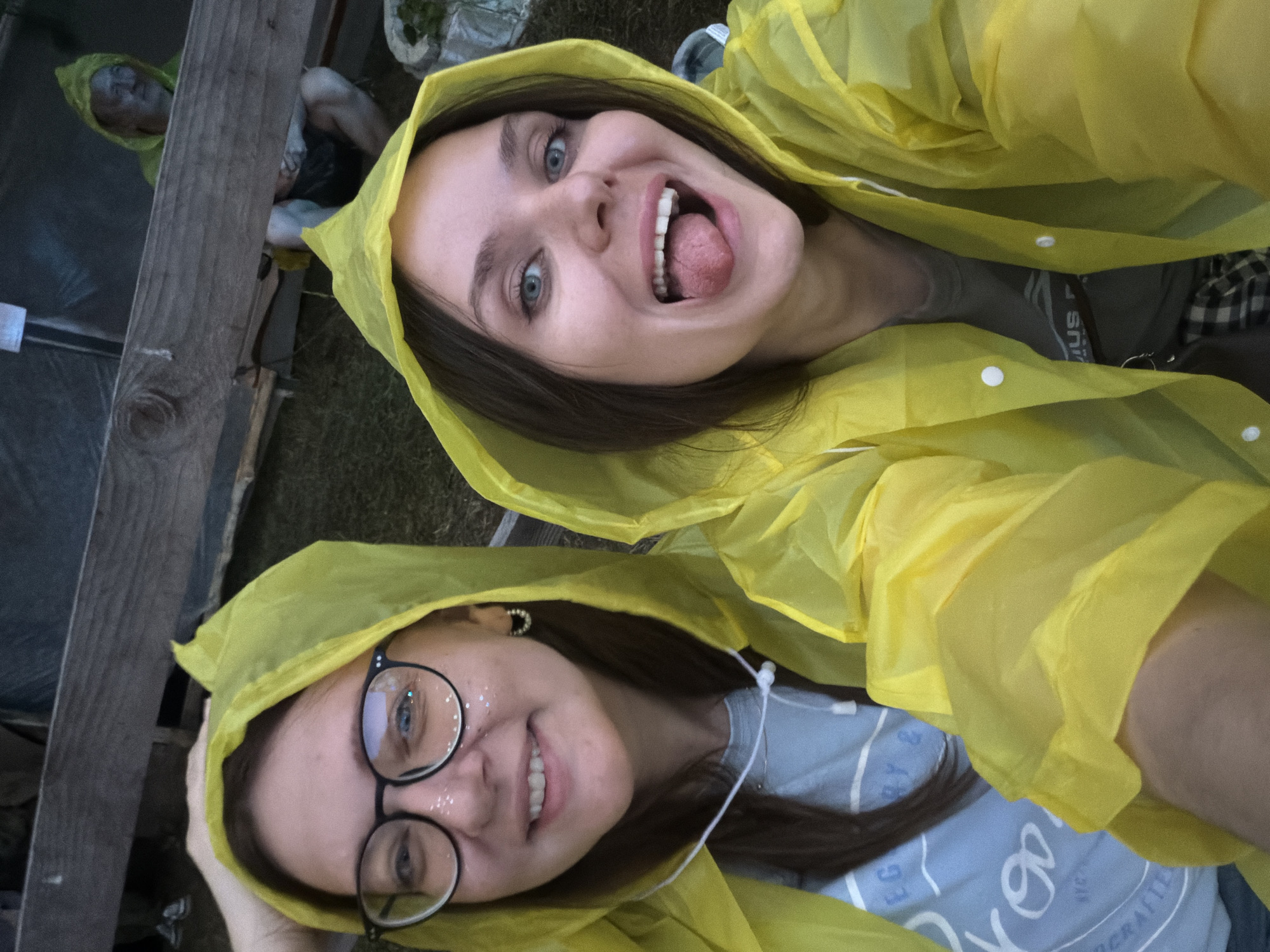 Two young women are pictured in close-up, wearing bright yellow hooded raincoats. The woman on the left wears glasses with round frames and has glitter applied to her cheeks. The woman on the right has her tongue sticking out. They appear to be outdoors, with a wooden structure and grass visible in the background. It is unclear if the location is in Il'inskiy Pogost, Russia.
