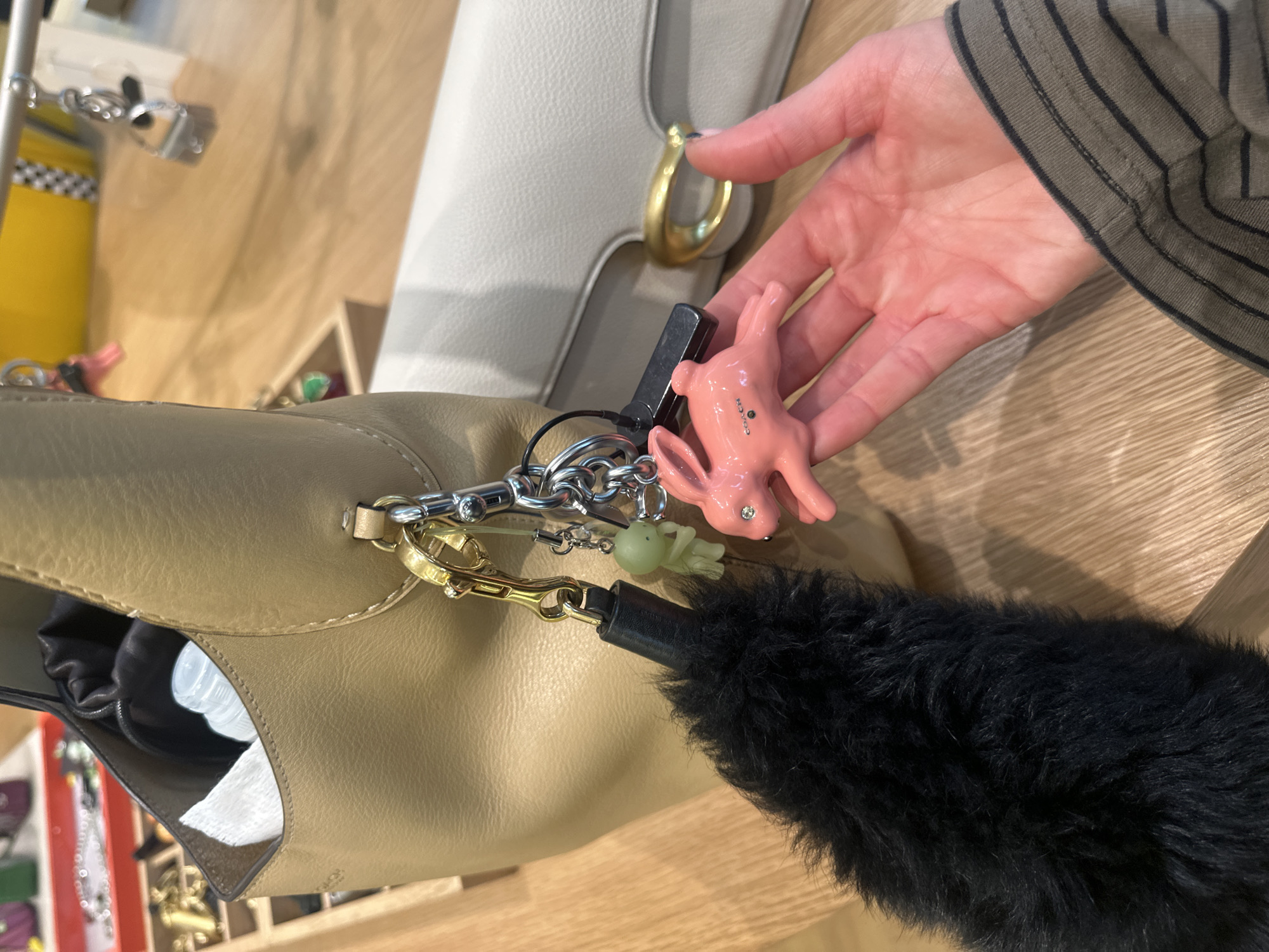 A close-up shot features a beige leather handbag with a black furry pom-pom attached to its zipper pull, along with multiple keychains, including a pink rabbit, held in a hand. The location is suggested to be in the city of London, United Kingdom, though this detail is inferred from the context provided and not explicitly visible in the image. The hand is lightly-skinned and belongs to a person wearing a shirt with black stripes on green/khaki fabric. In addition to the handbag, the image includes a white bag with a gold clasp visible in the background. A shelf below the bags holds additional bags and other objects. The background also shows a light-colored wood surface.