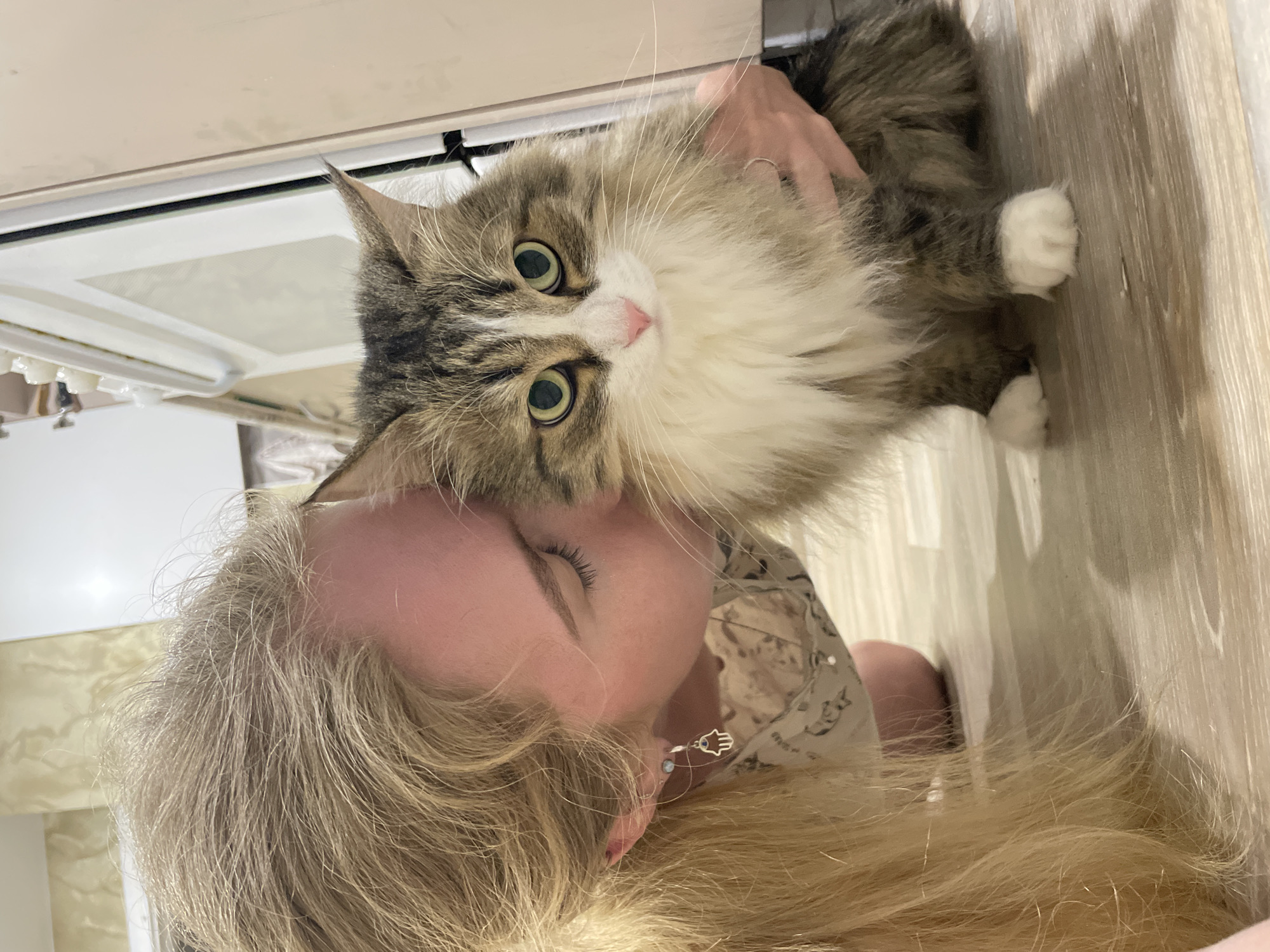 A person with long blonde hair is holding a brown and white long-haired cat. The cat is looking at the camera with green eyes. The background appears to be indoors, potentially in a home with light-colored flooring and a partial view of what seems to be a bathroom or bedroom. There is no evidence in the image to suggest it was taken in Izhevsk, Russia.