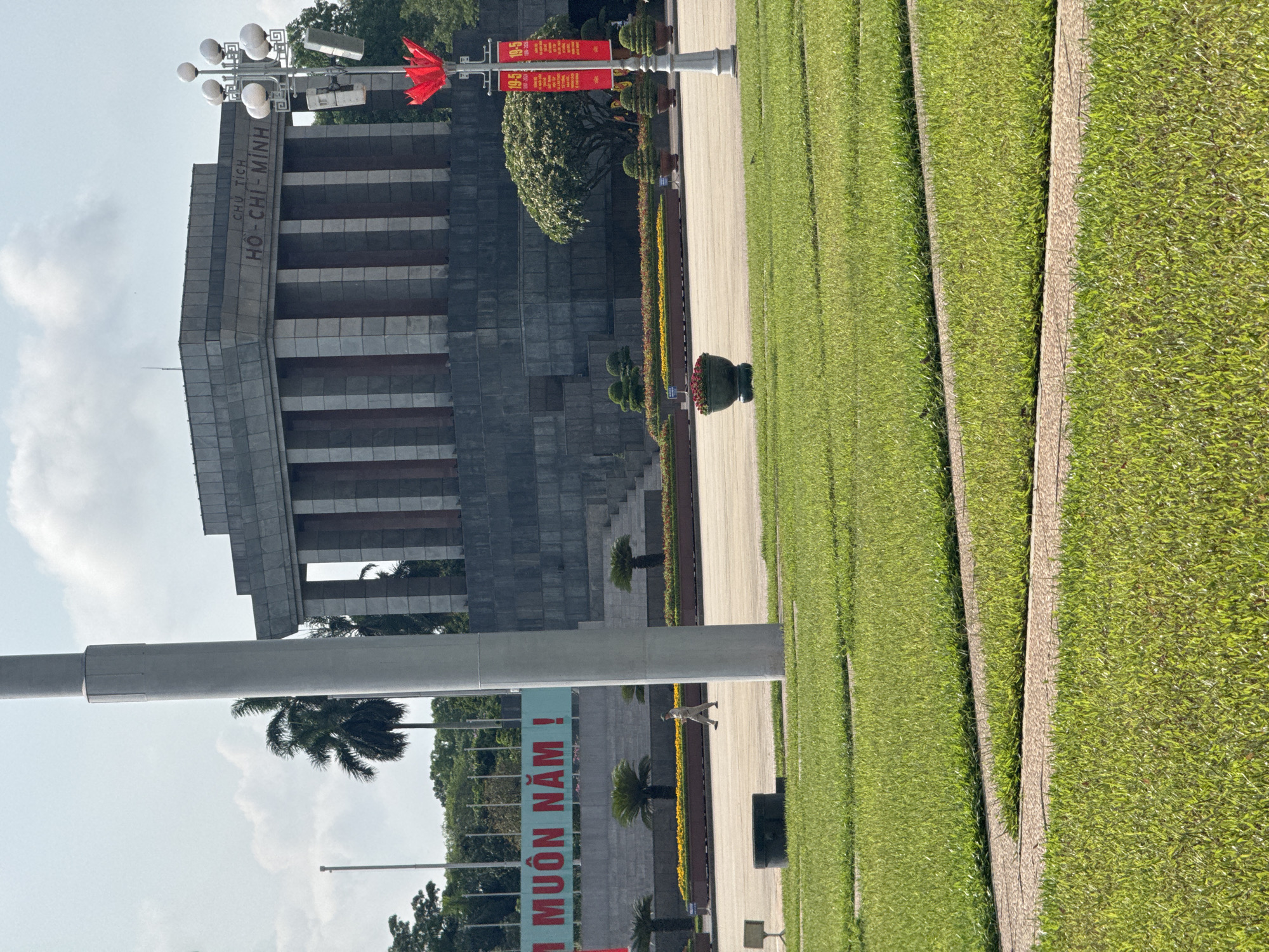 The image depicts the Ho Chi Minh Mausoleum in Hà Nội, Vietnam. The mausoleum is a large, gray stone building with the inscription "CHU TICH HO-CHI-MINH" on the front. A red flag is positioned on a light post, while other red banners are hanging on another post. A well-manicured lawn stretches in front of the mausoleum, with pathways and rows of small trees and flower arrangements bordering it. A uniformed guard is walking on the pathway. Banners with Vietnamese script are visible in the foreground. The sky is partly cloudy.