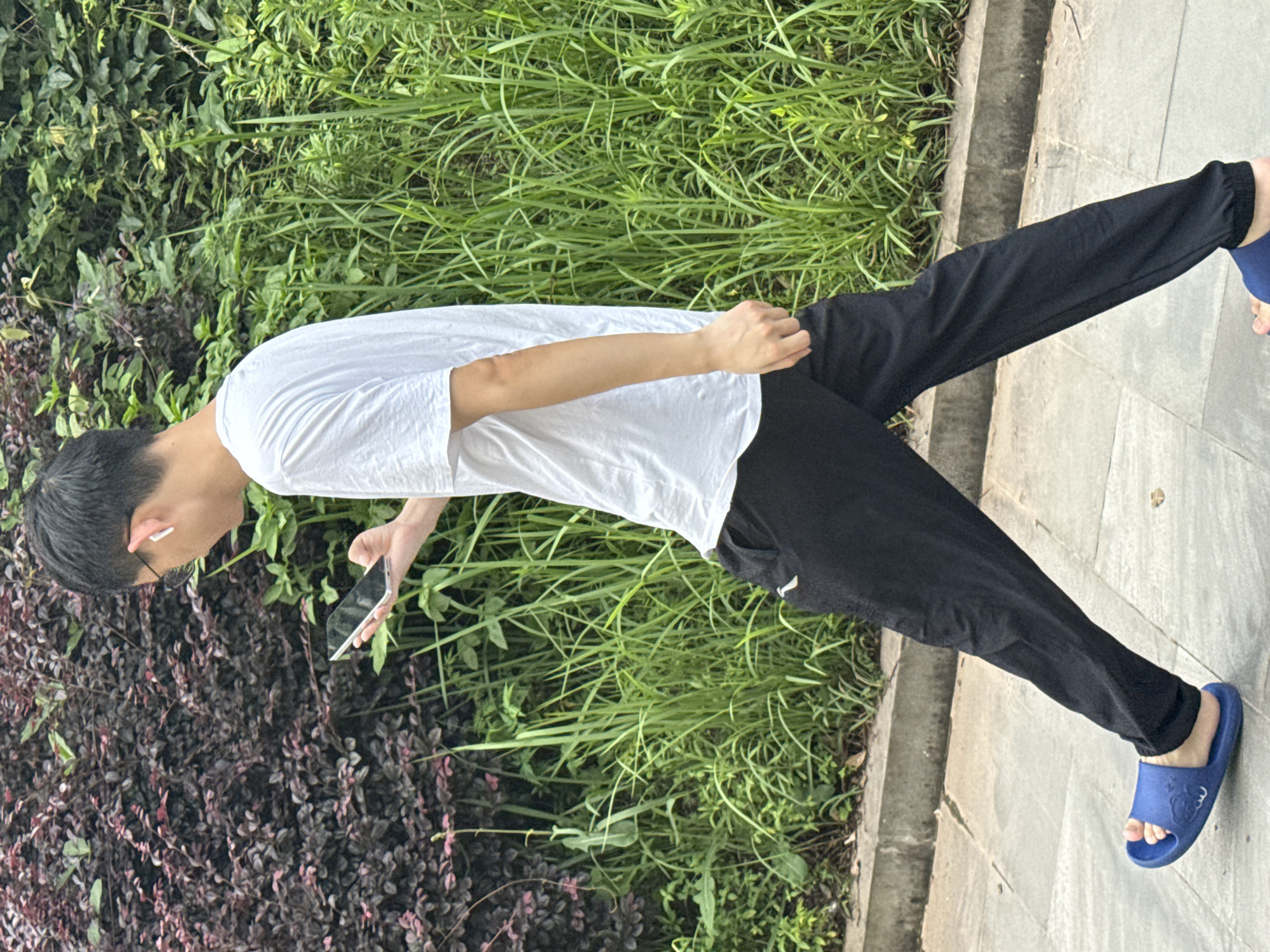 A young East Asian male, wearing a white t-shirt, black pants, and blue slides, walks along a tiled path in front of dense green foliage and a dark red-leaved shrubbery. He holds a smartphone in his right hand and wears white earbuds. The scene appears to be outdoors in an urban park or walkway in Jiangjin, China.