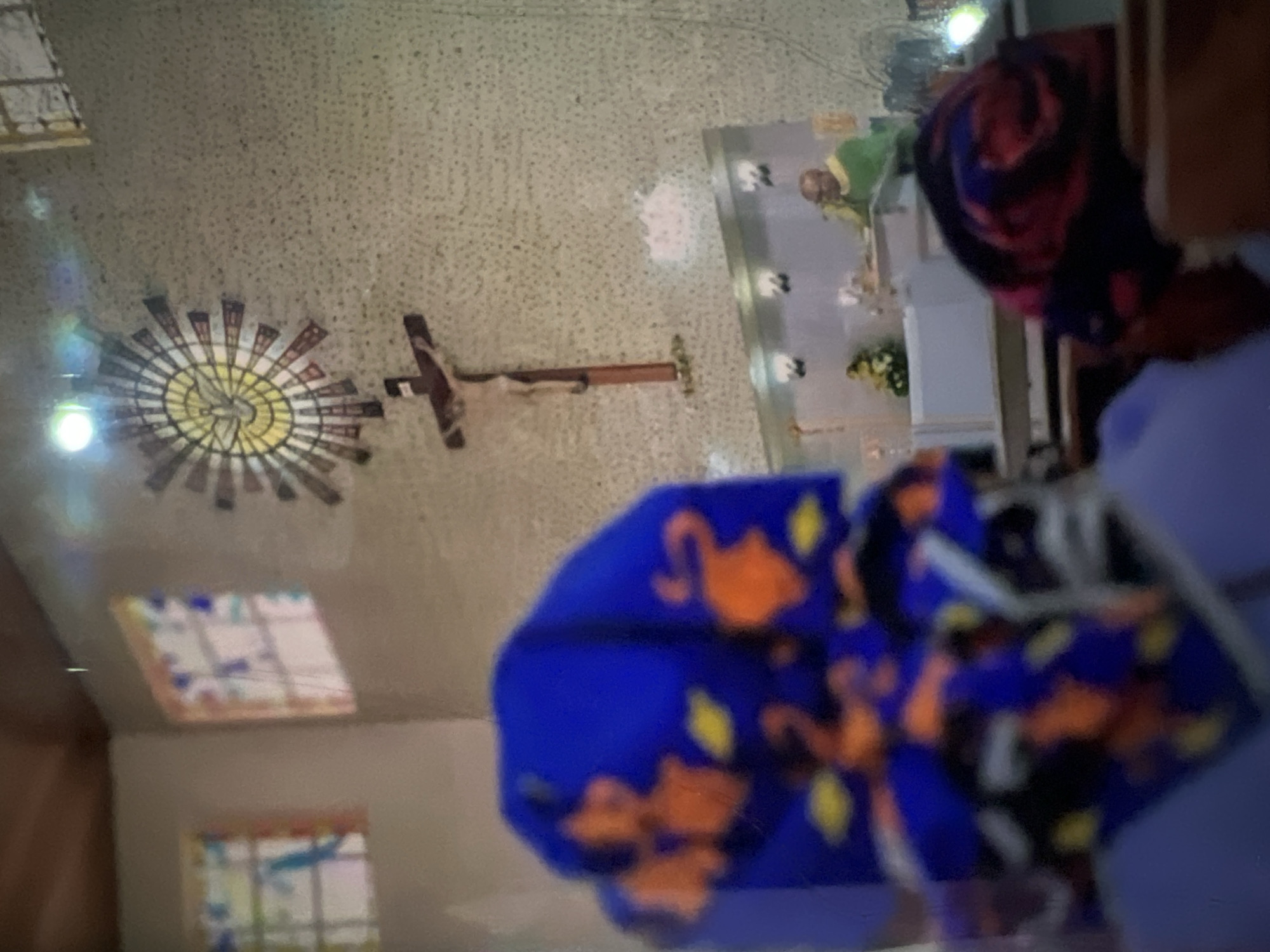 The image captures an interior scene likely within a church in Lagos, Nigeria. In the foreground, two individuals are partially visible, wearing vibrant headwraps with blue and orange patterns. The one on the left has a blue headwrap adorned with orange elephant motifs and yellow dots. The one on the right has a red and blue swirled headwrap.
The background reveals a religious setting. A crucifix hangs prominently on a textured wall. To the left of the crucifix, there are stained-glass windows with geometric patterns. Further back, a priest, wearing a green vestment, stands at a lectern, suggesting a religious service is in progress. The environment appears to be a place of worship, characterized by religious iconography and seating arrangements typical of a church.