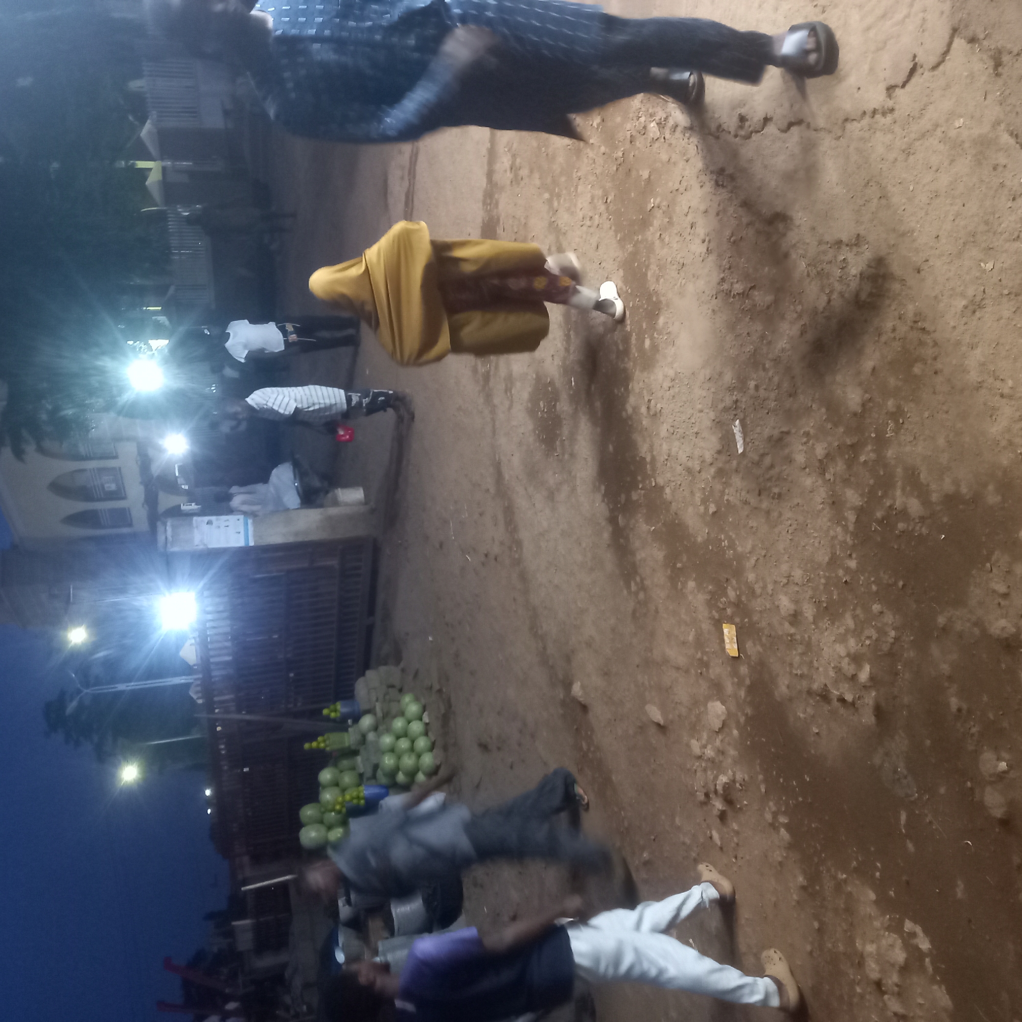 This image captures a vibrant, unpaved street or market scene in Barnawa, Nigeria, during the evening or night. The sky is dark blue, and the area is illuminated by several bright, artificial lights causing significant glare. The ground is a light brown, uneven dirt or sand, with scattered debris and textured patches.
Numerous people are visible, many blurred due to motion, indicating activity. In the foreground, one individual is seen wearing dark patterned clothing and sandals. Another person, seen from behind, wears a distinctive yellow-ochre garment over patterned trousers and light-colored shoes. Further into the scene, a group of people are gathered, with one wearing a white shirt and another a striped shirt. On the left side of the image, near a prominent market stall, individuals are also present; one wears a gray top and dark trousers, while another wears a purple top and white trousers with light brown shoes.
The most notable object is a large pile of green, spherical fruits, likely watermelons or large gourds, displayed on the ground or a low stand, with smaller yellow fruits mixed in. This arrangement suggests a fruit vendor's stall. In the background, several buildings are visible, including one with distinctive arched window-like features and a dark, possibly metal, gate or fence. The overall impression is a bustling outdoor market or street area active after daylight hours. No readable text is discernible in the image.