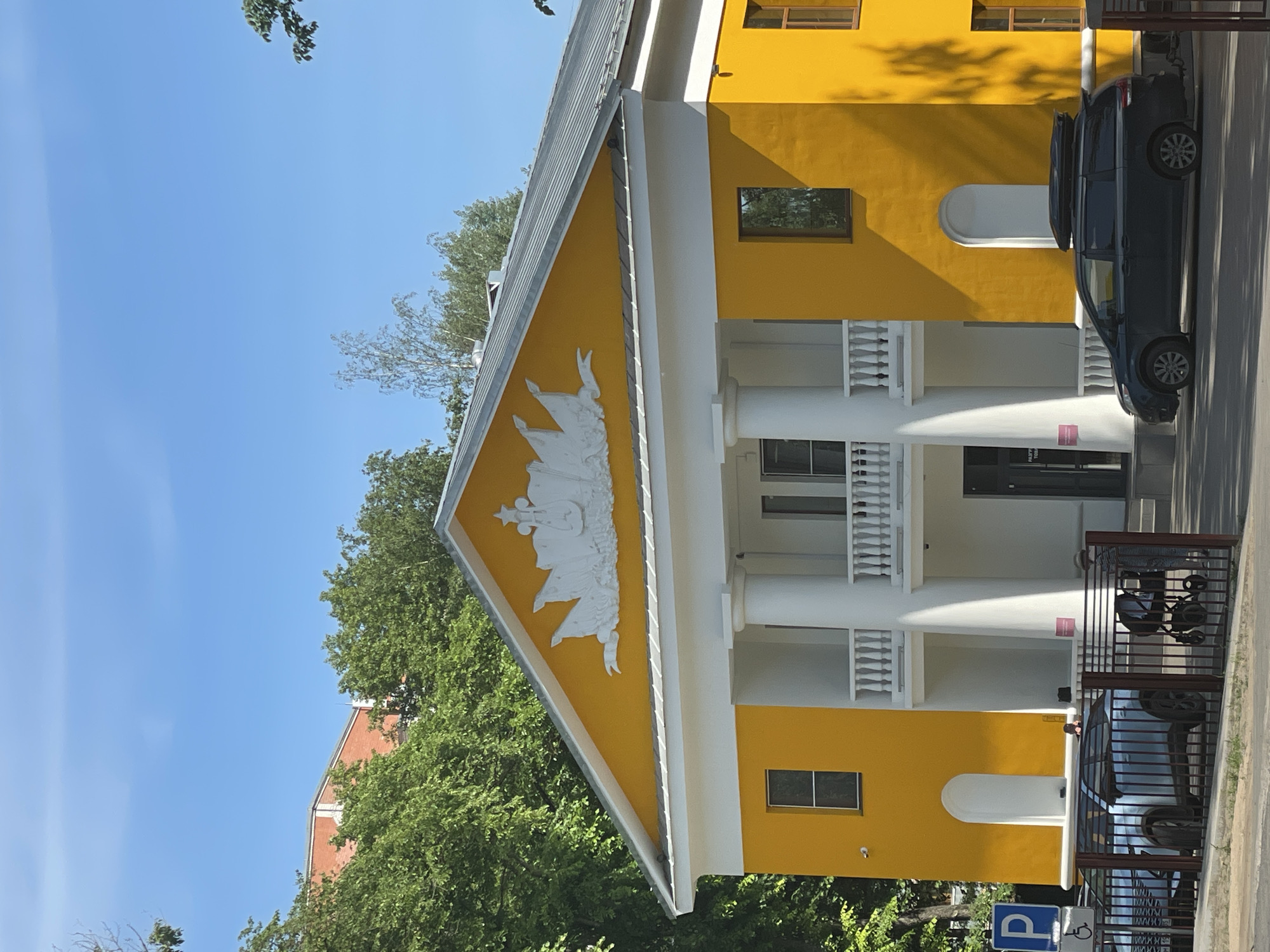 The image shows a bright yellow building with white columns and decorative elements, set against a clear blue sky and surrounded by lush green trees. The building has a classical facade with a triangular pediment featuring white relief ornamentation. There are two stories visible, with several windows and a balcony on the upper level. In front of the building, there is a dark-colored minivan with a roof box and a lighter-colored SUV parked behind a metal fence. A baby stroller is also visible near the stroller. A blue parking sign with a white "P" is present on the left side of the image. This building is located at 15 Sovetsky Prospekt in Vologda, Russia, which is the former Vologda Theological Seminary, currently housing the Vologda Regional Picture Gallery.