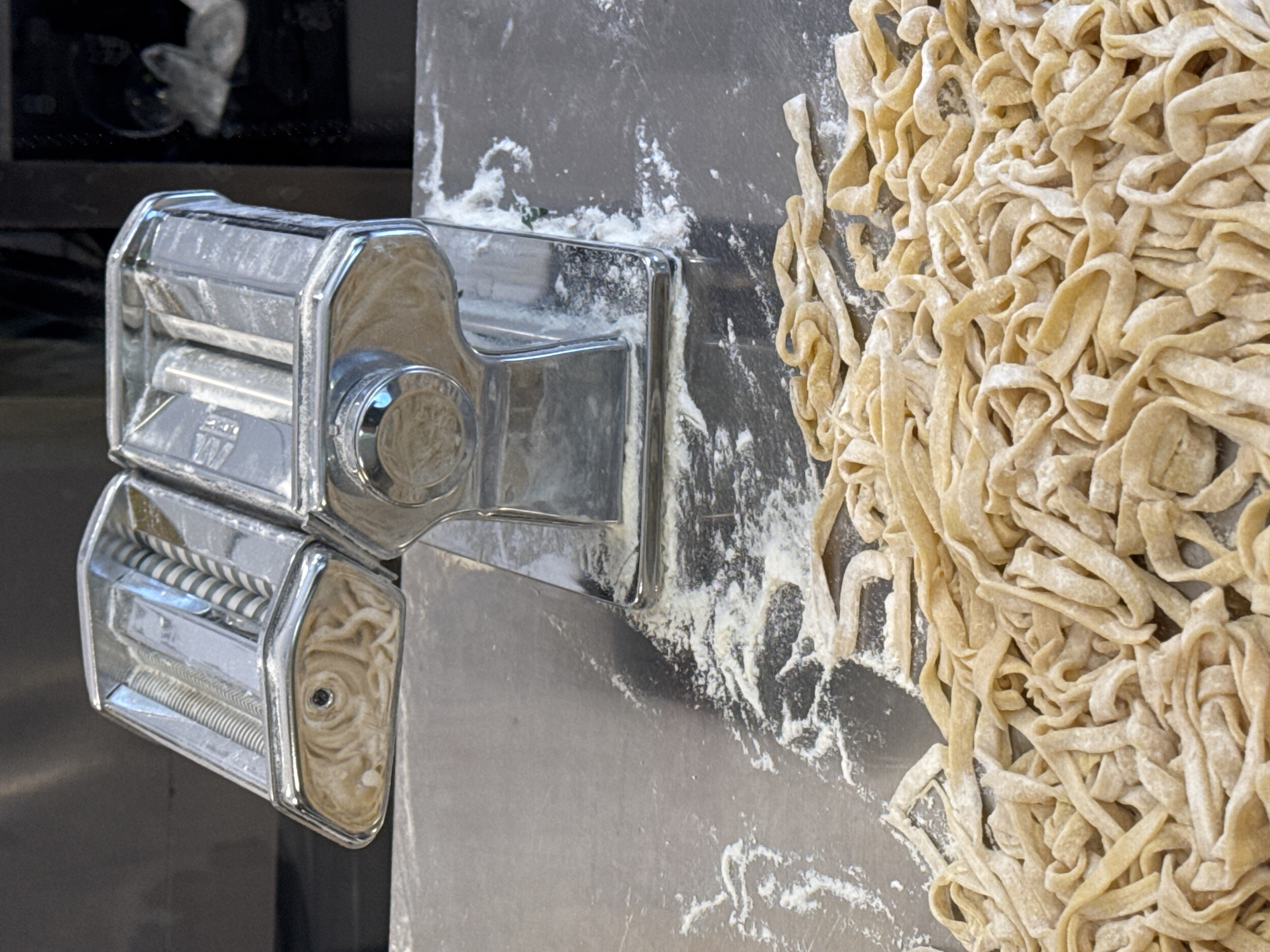 The image displays a close-up view of a pasta preparation scene within what appears to be a kitchen or food preparation area in Budva, Montenegro. A chrome-plated manual pasta machine is positioned in the upper center of the frame, visibly coated with a fine layer of white flour, indicating recent activity. The machine's internal mechanisms, including its rollers, are partially visible through openings, also dusted with flour. Below and extending into the foreground, a large quantity of freshly cut, light-yellow pasta noodles, consistent with tagliatelle or fettuccine, is piled on a reflective, grey metallic surface, likely stainless steel. These noodles are also lightly floured to prevent sticking. Scattered white flour residue is present on the metallic surface around the base of the pasta machine and amidst the fresh pasta. The background is largely out of focus and dark; an indistinct, blurred shape, possibly a reflection or a distant object, can be observed on the right side against a darker backdrop, while the left side of the background is a uniform dark grey. The scene is illuminated by an overhead light source, casting reflections on the metallic surfaces.