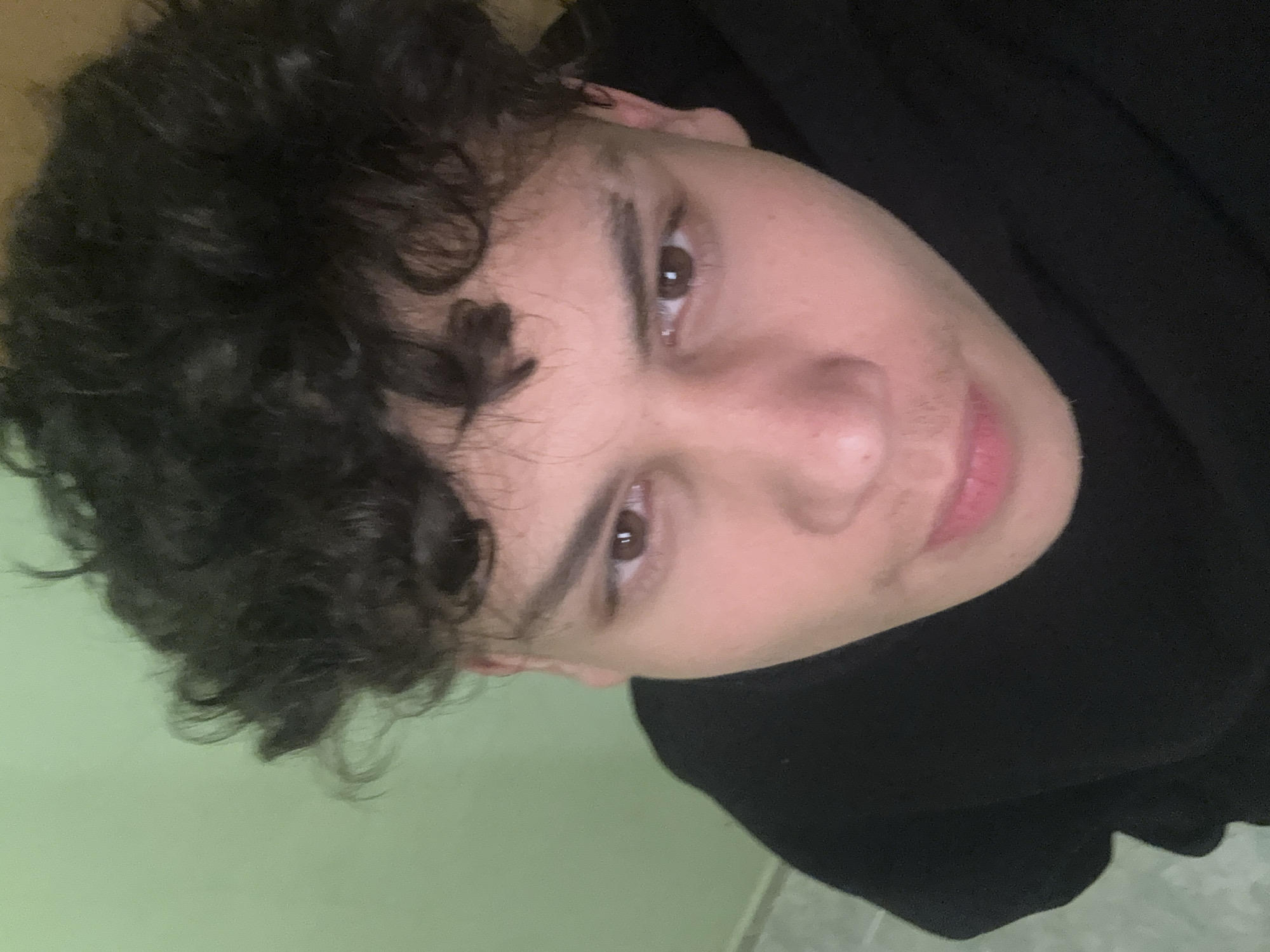 A close-up, portrait-style shot from an upward angle features a person with dark, curly hair. The individual has light skin, brown eyes, and defined eyebrows. A small amount of facial hair is visible above the upper lip. The person is wearing a black garment, likely a hooded sweatshirt or jacket. The background appears to be a light green or pale wall. The setting or location of this image in the city of Sofia, Bulgaria, is not discernable.