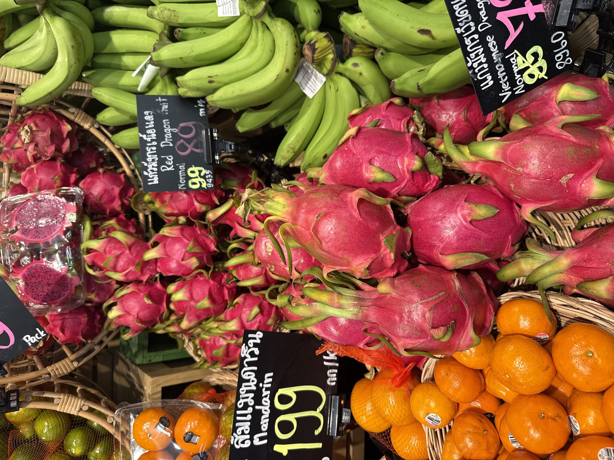 The image shows a close-up of a fruit stall, likely in an outdoor market or a grocery store, featuring a variety of fruits. In the foreground, a basket overflows with bright orange mandarins, some of which have stickers with a circular logo. To the right and above the mandarins are vibrant pink dragon fruits, some whole and some sliced, revealing their speckled interior. Behind the dragon fruits, bunches of green bananas are visible. A black sign with white and yellow text displays pricing information and fruit names in both Thai and English, including "Mandarin" priced at "199" and "Red Dragon Fruit" priced at "99". Another sign indicates "Vietnamese Dragon Fruit". The overall impression is of a colorful and abundant display of fresh produce. The context of Pattaya City, Thailand, suggests this scene is part of the city's vibrant food scene, possibly at a local market or a supermarket's produce section.