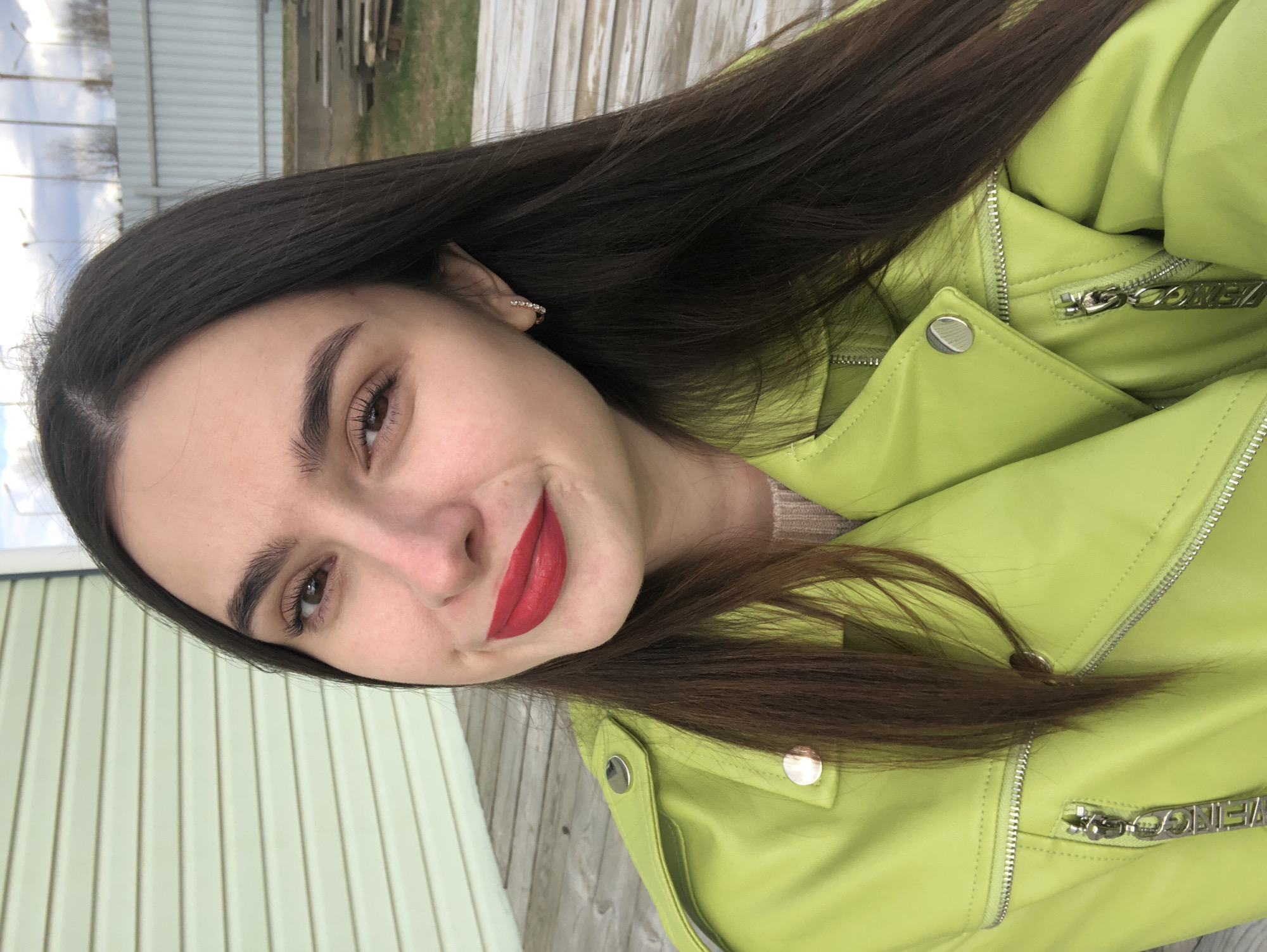 A young woman with long, dark brown hair and fair skin is depicted in an outdoor portrait, smiling gently at the camera. Her hair is parted to one side and flows over her shoulders. She wears vibrant red lipstick and has well-groomed, dark eyebrows and visible eyelashes framing her dark brown eyes. A small, sparkling hoop earring is visible in her right ear.
She is dressed in a striking lime green (or chartreuse) leather-style moto jacket, which features multiple silver zippers and prominent silver snap buttons on the lapels and shoulders. A light beige or cream-colored knit top is visible underneath the jacket at the neckline. One of the jacket's chunky silver zipper pulls has the word "COMEZI" faintly engraved on it.
The background indicates an outdoor, daytime setting. To her left, there is a light green building or wall with vertical paneling. To her right, a light-colored, weathered wooden structure, possibly a fence or deck, is visible. In the distant background, a cloudy sky and hints of utility poles or trees can be seen. The lighting is soft and even, suggesting either overcast conditions or indirect sunlight. The woman's expression is pleasant and relaxed.