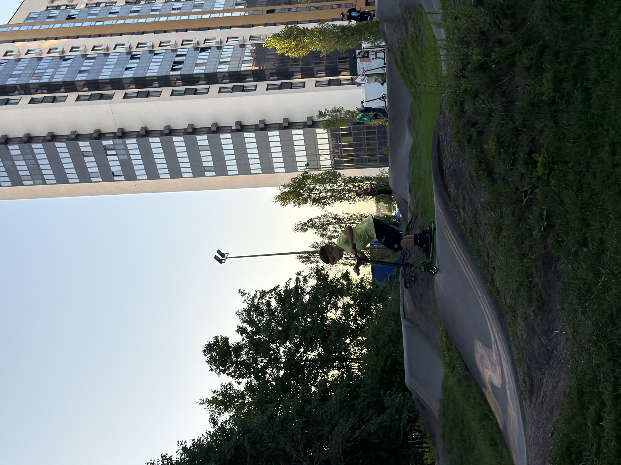 In the city of Unknown, Unknown, a boy on a green scooter navigates a dirt bike track. He wears a light green shirt and black shorts with knee pads. Behind him, other children are seen on scooters, and a tall, modern building with many windows stands in the background. Trees and a lamppost are also visible. The sky is clear and pale.