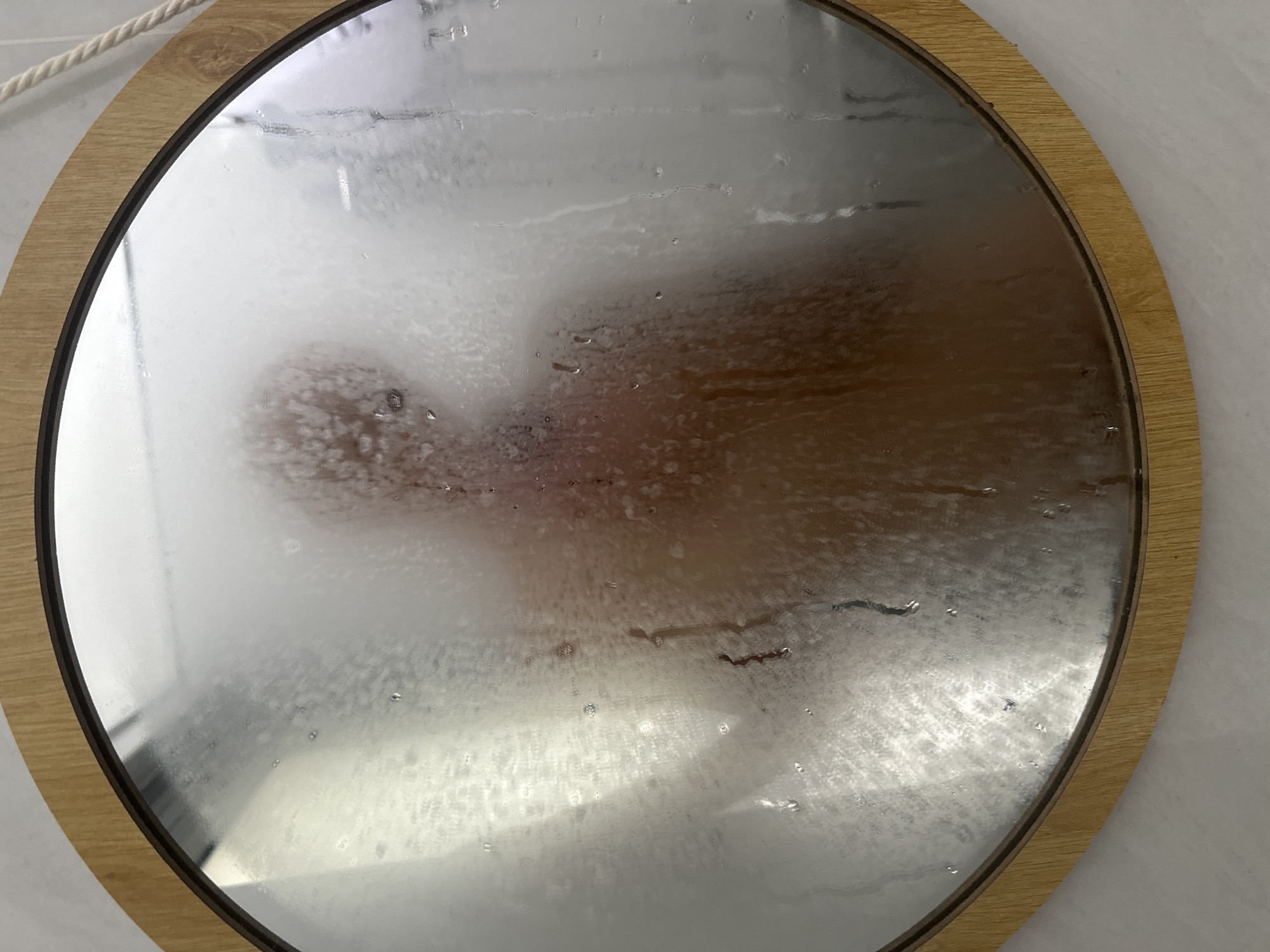 A circular mirror with a wooden frame is heavily fogged with condensation, obscuring the reflection of a person. The mirror features a light brown wooden frame with a thin black inner border. Water droplets and streaks are visible on the glass surface, indicating high humidity, likely from a shower.
The central subject is a blurry, indistinct human silhouette reflected in the steamed-up mirror. A head-like shape and torso are vaguely discernible, suggesting a person facing the mirror. The clarity of the reflection varies across the surface, with some areas more opaque due to thicker steam, and others showing faint details through the condensation.
In the top-left corner, outside the mirror's frame, a white braided rope or cord is visible against a light-colored wall. The scene evokes the aftermath of a warm shower, creating a hazy and intimate atmosphere. No text is visible in the image. The location context for this image is Mũi Né, Vietnam.