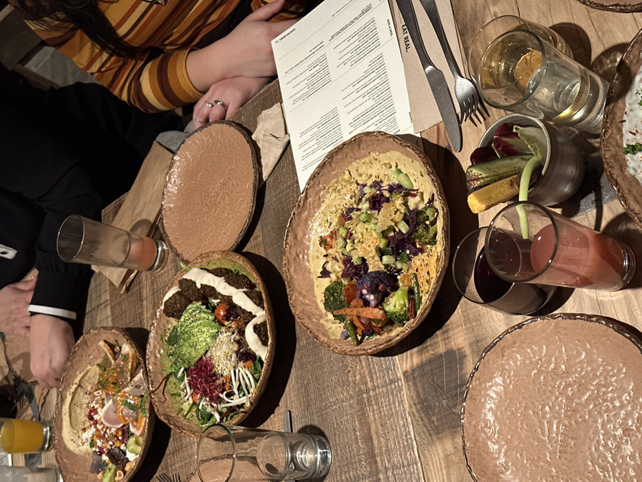 A wooden table displays an array of dishes, glasses, and dining accessories. Bowls filled with salads, vegetables, and hummus are accompanied by empty plates. Glasses of juice and water are scattered around. Cutlery rests next to a menu card with the words "EAT REAL." People wearing a striped sweater and a black jacket are seated at the table, engaged in the meal. The overall ambiance suggests a casual dining setting.