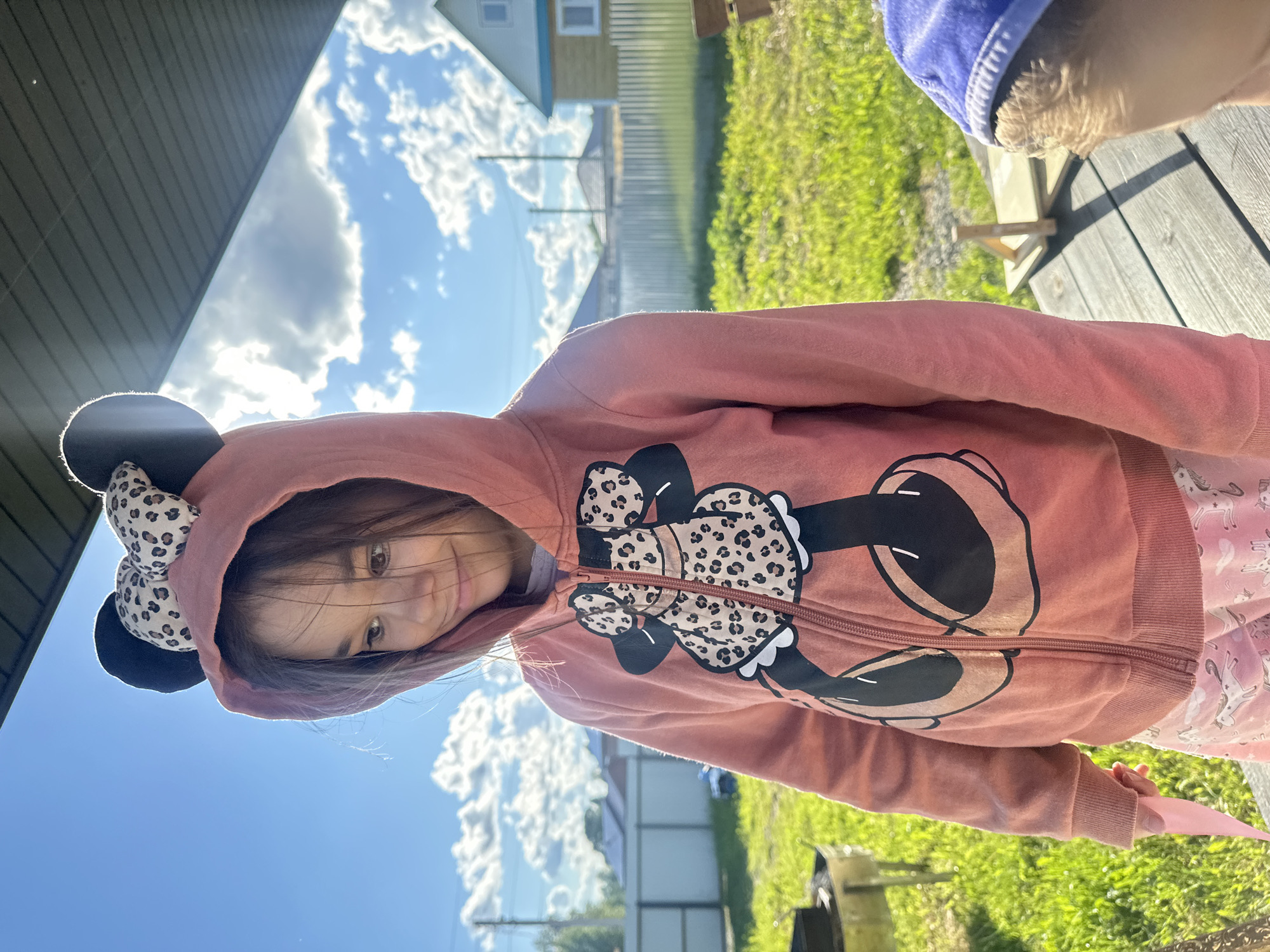 A young girl in a pink Minnie Mouse hooded jacket is outdoors. The background shows green grass and a partly cloudy sky. A building is also visible. The location is Kharnavy, Russia.