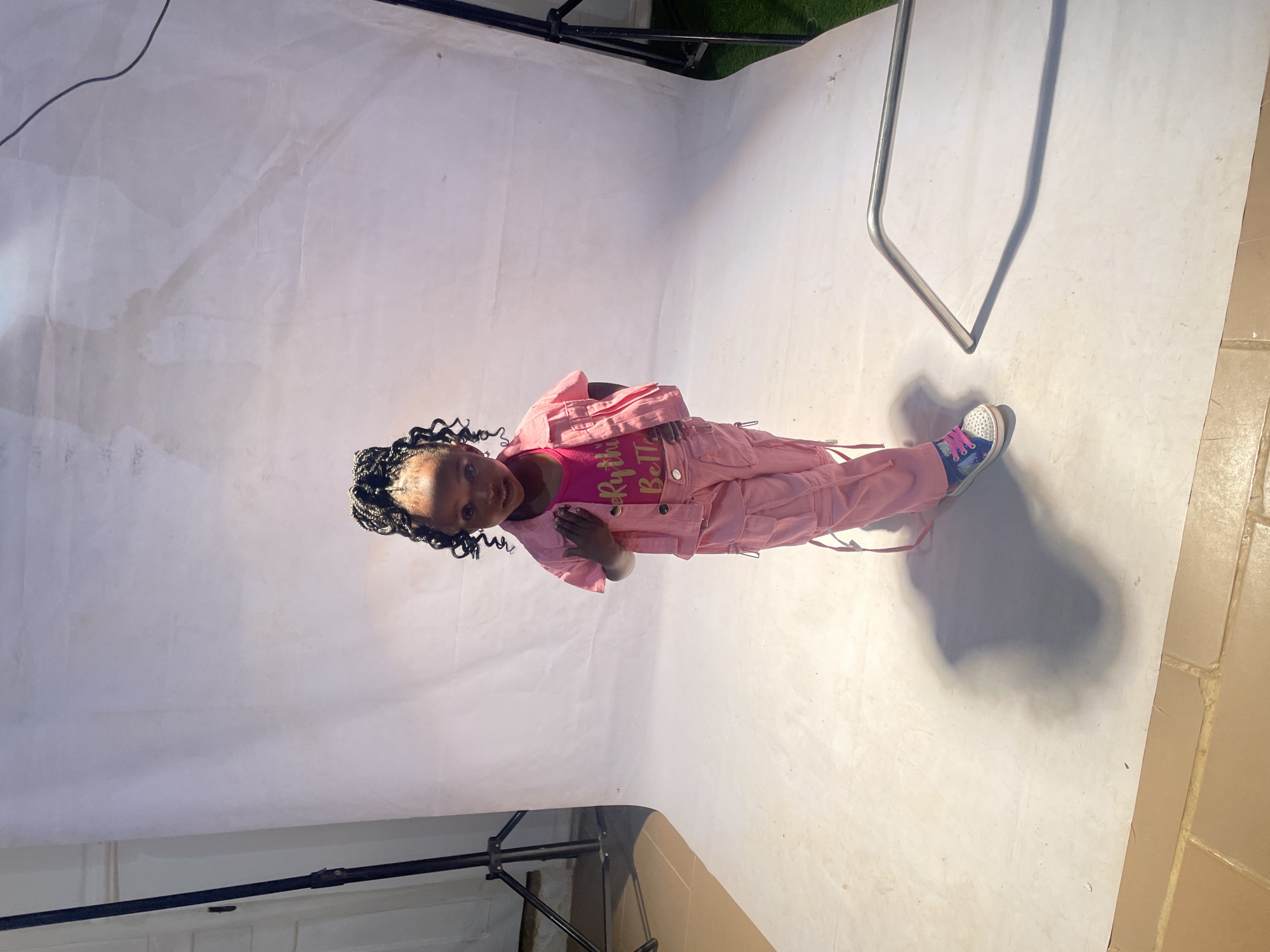 A young girl stands in what appears to be a studio setting, possibly located in Jos, Nigeria, although no definitive location identifiers are present. The girl, with dark hair styled in braids, is wearing a pink outfit consisting of a pink top with text and a matching jacket and pants. She is also wearing blue and white sneakers. She stands on a white backdrop that curves to simulate a continuous background. Lighting equipment is partially visible, including a light stand on the left and a metal frame on the right. The floor in the lower right corner transitions to a tiled surface. The girl is looking at the camera with a slight smile.