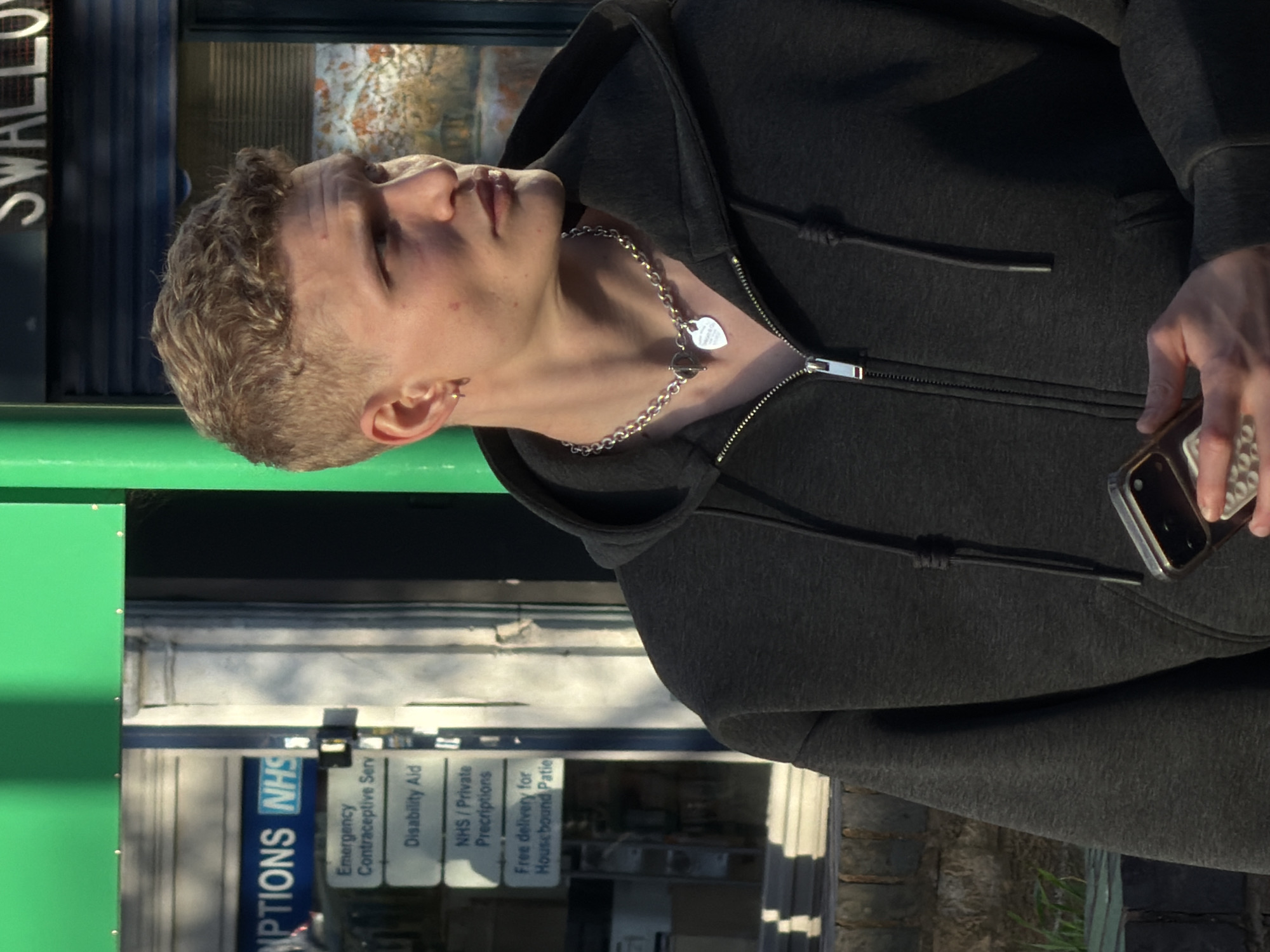 The image shows a fair-skinned individual wearing a dark grey zip-up hooded sweatshirt and a silver chain necklace with a heart-shaped pendant. They are holding a smartphone. The background includes signage indicating a pharmacy in London, United Kingdom, featuring NHS (National Health Service) branding. Signs are visible with information about services such as emergency contraceptive services, disability aid, NHS/Private prescriptions, and free delivery for housebound patients. There is also a partial view of a street sign with the word "SWALLOW" visible. The image is rotated counter-clockwise.