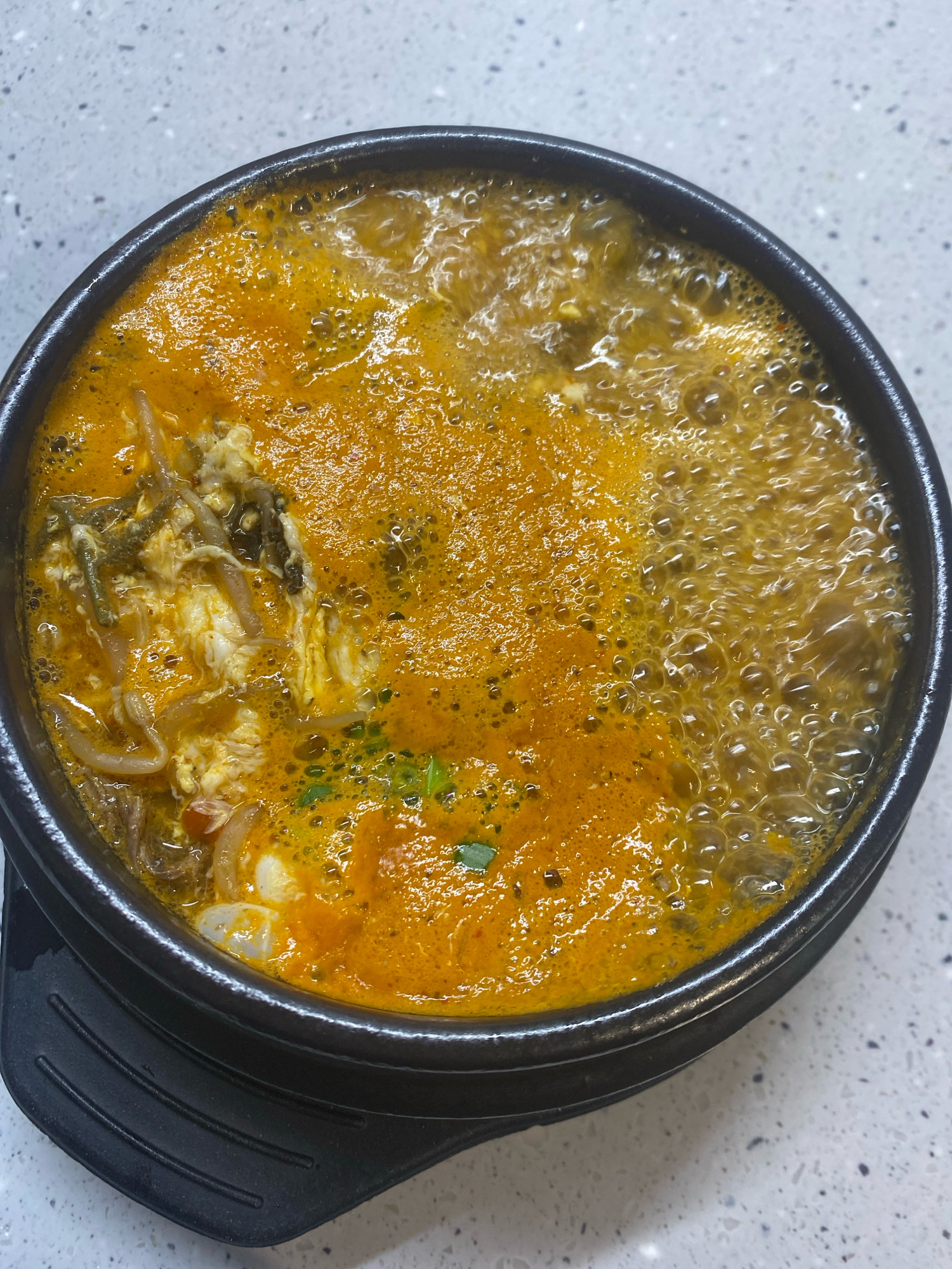 A top-down, close-up shot shows a steaming bowl of Korean sundubu-jjigae (soft tofu stew) on a speckled white countertop. The stew is a vibrant orange-red color, with visible chunks of soft tofu, wispy strands of egg, and other ingredients. The surface of the stew is actively bubbling, indicating it is hot and recently prepared. The black ceramic bowl has a textured outer ring and sits on a black insulated base. The environment appears to be an indoor setting, likely a kitchen or dining area, with no visible city or geographical markers to suggest a location in Astana, Kazakhstan.