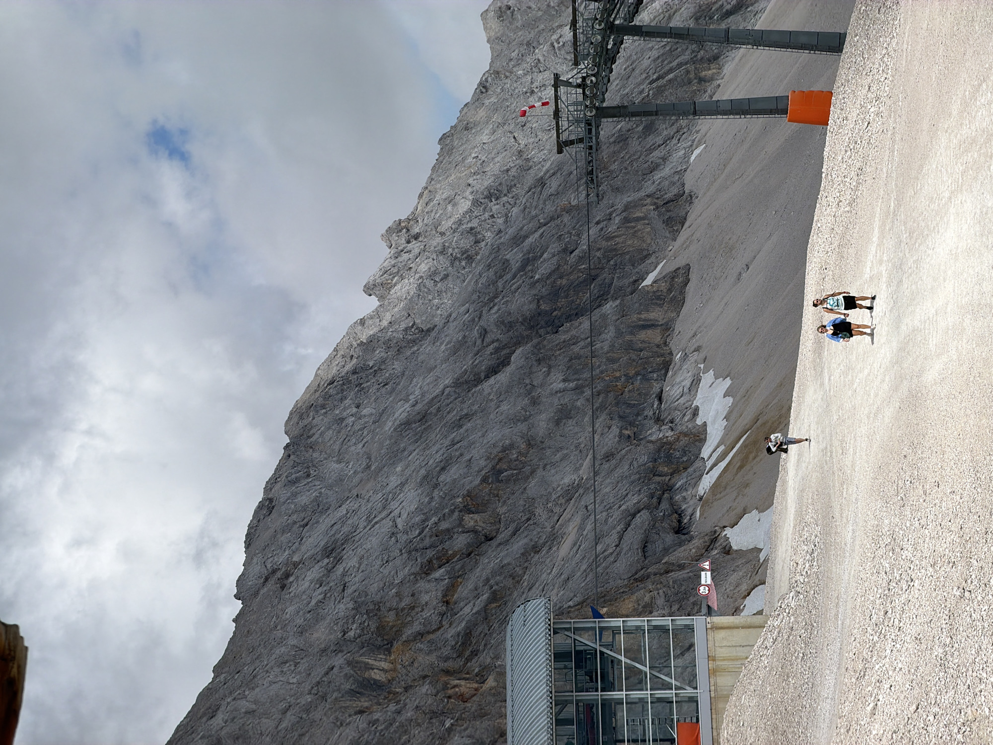 The image depicts a mountainous landscape near Garmisch-Partenkirchen, Germany, likely at a high altitude, suggested by the presence of snow patches on the slopes and the cable car infrastructure. In the foreground, a gravel path winds through the scene. Two individuals, a man and a woman, are walking towards the right on this path. The man wears a white tank top and dark shorts, while the woman is dressed in a blue jacket over a dark outfit. Further to the left and further back, another person, also appearing to be a woman, walks towards the left, carrying a dark bag.
To the left of the path is a modern building with a metal roof and large glass windows, possibly a cable car station. A sign with a circle and the number "3,50" is visible on the side of the building, along with a triangular warning sign. To the right, a large cable car support tower rises from the ground, with the characteristic red and white striped windsock attached to its upper structure. The background is dominated by a massive, rugged mountain face, characterized by steep inclines and rocky textures. The sky above is overcast with scattered clouds. The overall impression is a serene, outdoor mountain environment with signs of human activity and infrastructure related to tourism or transportation.
